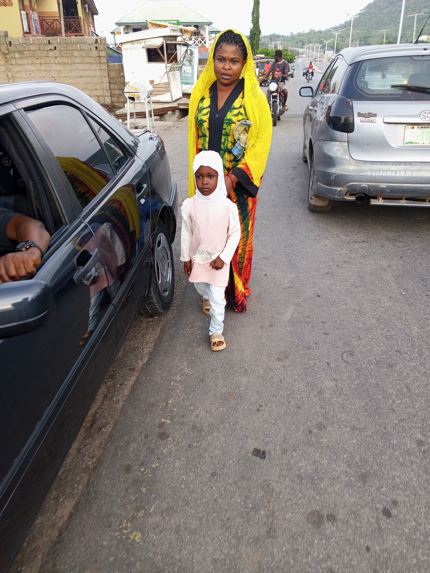 A full-shot image captures a woman and a young child walking on the side of a paved road in what appears to be a bustling area of Zango-ndaji, Nigeria. The woman, standing slightly behind the child, wears a bright yellow head covering and a long, multi-colored patterned dress. She has a neutral expression and holds a light-colored object in her left hand. The child, likely a girl, walks in front wearing a white head covering (hijab), a light pink long-sleeved top, light blue pants, and open-toed sandals. She looks directly at the camera with a serious expression.
To their left, a dark-colored sedan is partially visible, with a person's arm and hand, wearing a watch, seen through the driver's side window. Reflections of the woman and child are visible on the car's body. Further down the road on the right, a silver Toyota hatchback, identifiable by "TOYOTA" badging and a "LAGOS" license plate (partially visible, including "178"), is either parked or moving slowly. Two motorcyclists and other pedestrians are visible in the distance on the road.
The background features various buildings, including informal structures and shops. One small booth clearly displays "POS SERVICES" and another sign reads "MAMA LYD..." with "OMAR FOODS" visible on a white stand. Distant hills or mountains frame the horizon. The time of day appears to be late afternoon or early evening, characterized by soft, diffuse lighting, suggesting an overcast sky or approaching dusk. The overall scene depicts everyday life along a roadside in an urban or semi-urban Nigerian setting.