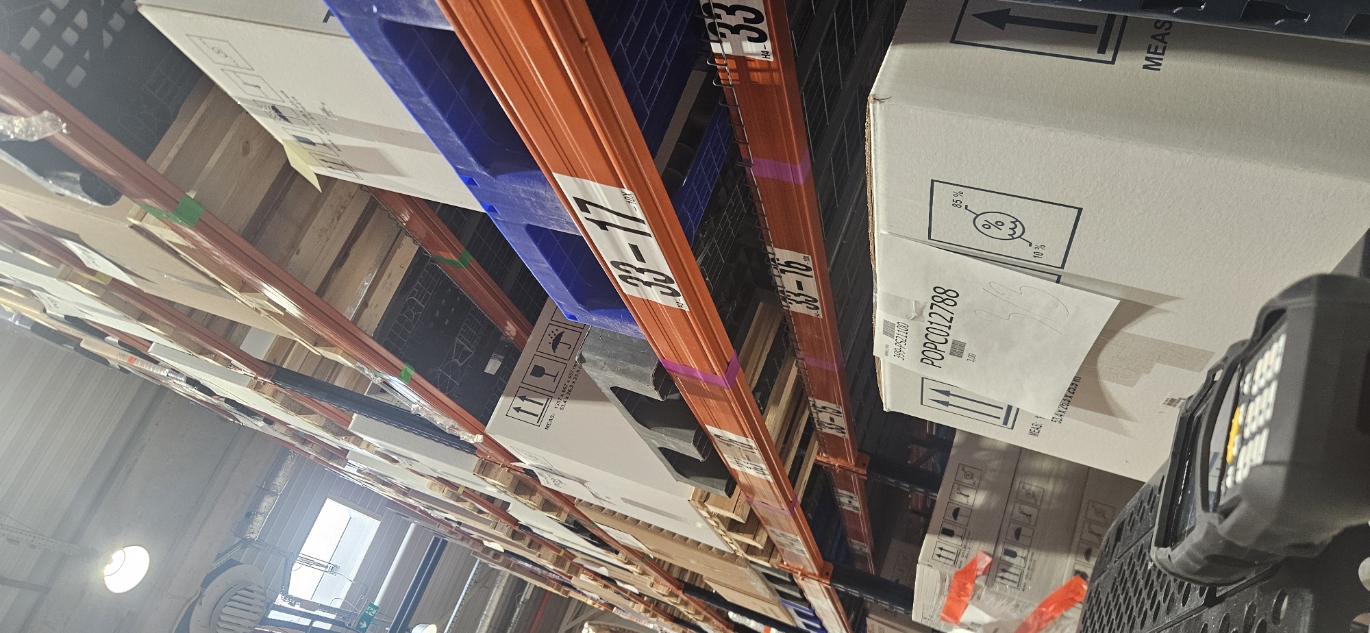The image depicts a warehouse storage system, likely located in Plovdiv, Bulgaria, though that is not definitively evident from the image. The scene shows multiple levels of shelving units filled with boxed items and blue plastic pallets. The metal shelving is painted orange and labeled with black and white alphanumeric codes, such as "33-17" and "33-16".
The boxes contain various symbols and text, including measurements and handling instructions (e.g., umbrella symbol indicating protection from rain). Some boxes are labeled with order numbers such as "POPC012788" and codes like "399-PS21100". There is also a barcode scanner in the foreground, suggesting inventory or logistics operations are taking place. The environment suggests a commercial or industrial storage facility.