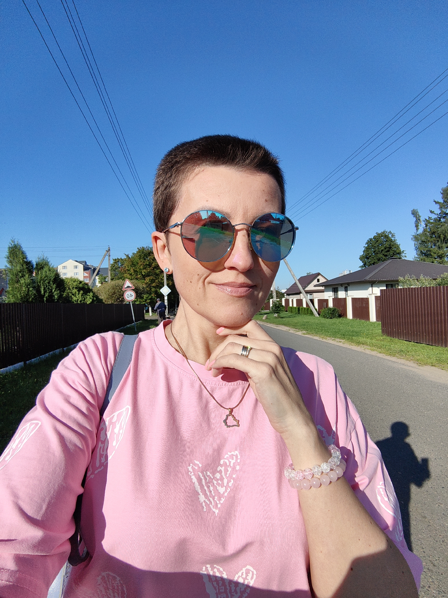 This is a medium shot, selfie-style photograph of a woman with short, dark hair and sunglasses, taken outdoors on a sunny day. She is wearing a pink sweatshirt with a white heart design on the front. Her right hand is resting on her chin, and she is wearing a ring on her index finger and a bracelet on her wrist. The background shows a paved road, houses with brown fences, trees, and a clear blue sky with power lines overhead. In the distance, there are traffic signs and a person riding a bicycle. The scene appears to be a residential street in Fanipal, Belarus.
