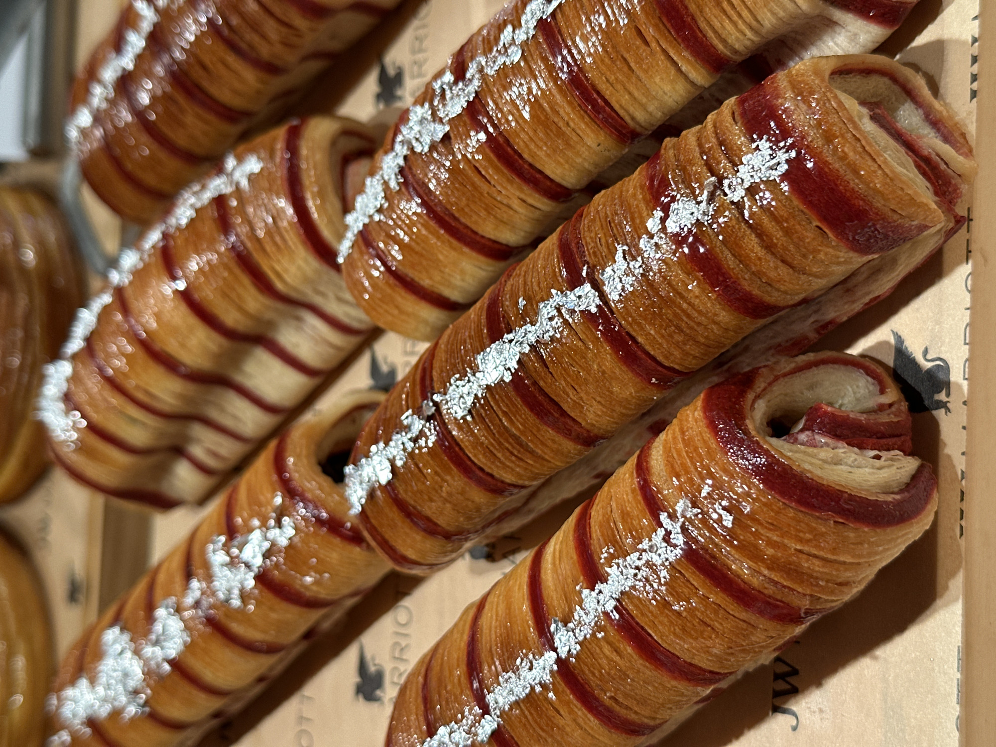 The image displays a close-up, overhead view of several pastries arranged on a tray. The pastries are elongated and appear to be made of layered dough, with a golden-brown exterior. They are adorned with bright red stripes running lengthwise and sprinkled with what appears to be edible silver flakes. The pastries are partially unfurled, revealing a creamy, reddish filling. In the background, hints of other pastries and a wooden surface are visible. The lighting is bright, highlighting the textures and colors of the baked goods. The scene is devoid of people and activity, focusing solely on the presentation of the food. The context of Vagator, India, is not discernible from the visual information in the image itself.