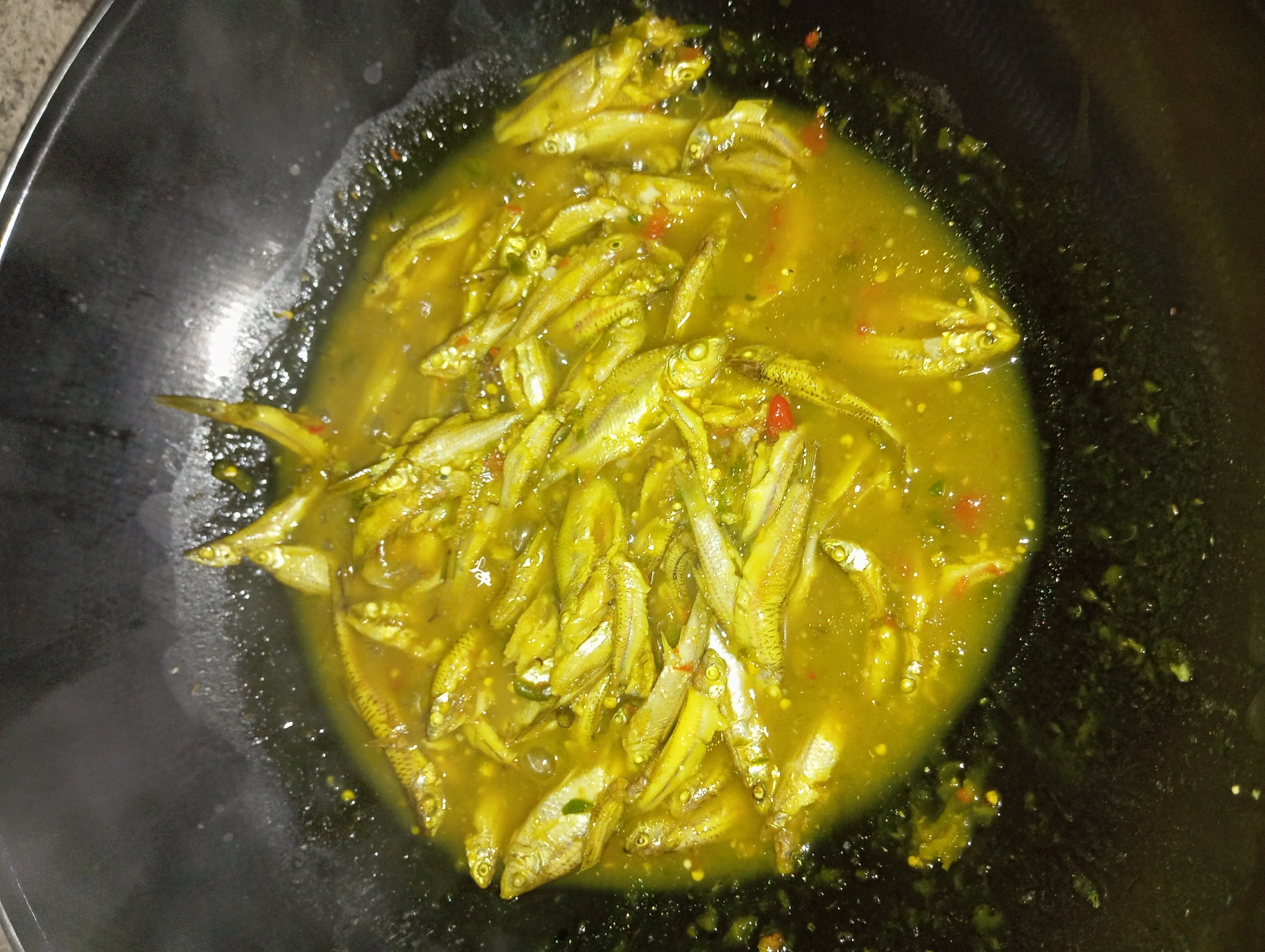 This is a close-up, overhead, forensic photograph of small, whole fish being cooked in a yellow, saucy broth within a dark, round cooking vessel, likely a wok. The fish appear to be a type of sardine or anchovy and are densely packed together. The broth contains visible red chili pieces and other unidentifiable spices. The cooking vessel has a slightly textured dark surface, and some splattering of the broth is evident on its inner rim. The lighting suggests an indoor setting with some ambient light, and the focus is entirely on the food being prepared. The image does not contain any elements that would place it specifically in Rejosari, Indonesia.