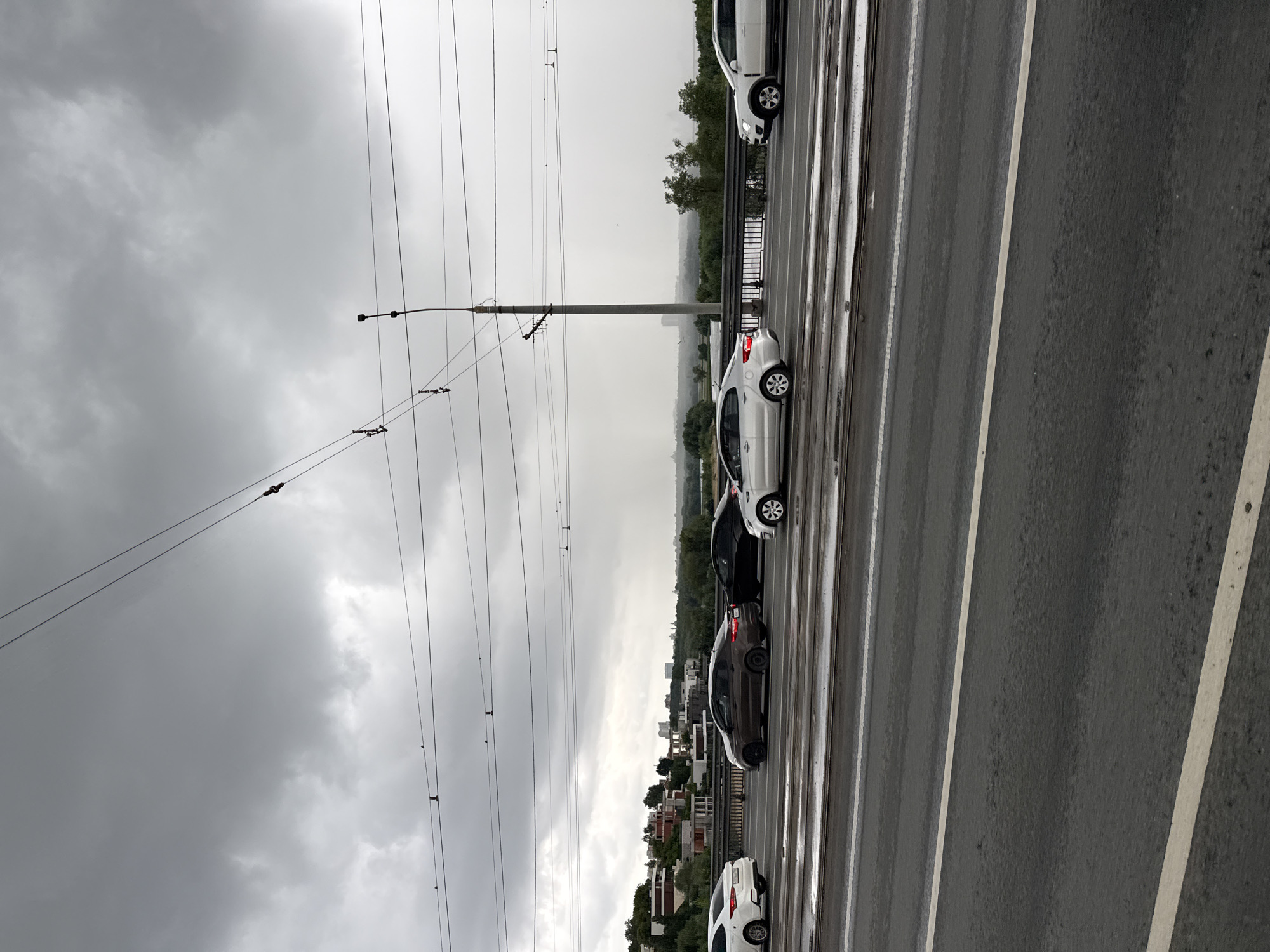 The image depicts a cloudy, overcast day with traffic on a bridge. Several cars, including a white sedan, a dark sedan, and a silver sedan, are visible on the bridge. The road appears wet, suggesting recent rain. In the background, there are buildings and trees, indicating an urban or suburban setting. The sky is filled with heavy, dark clouds, and there is a visible haze or light rain in the distance. Overhead, power lines and a traffic light pole are present, typical infrastructure for a city. This scene is consistent with the view from the Chistopolskaya bridge in Kazan, Russia, which spans the Kazanka River and connects to the city's infrastructure, often experiencing this type of weather.