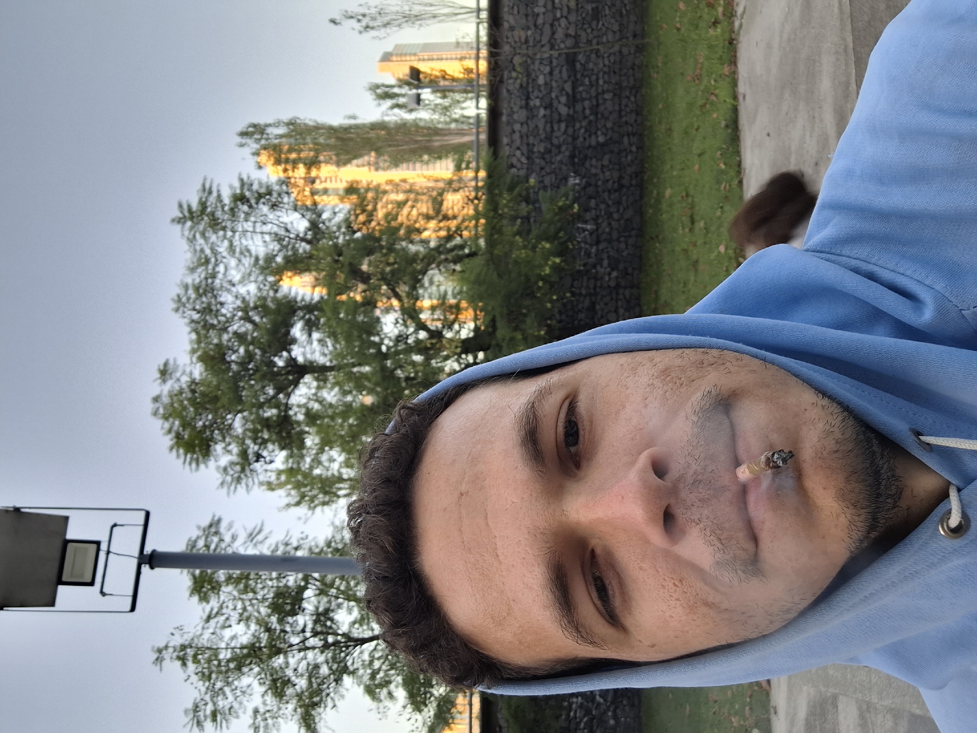 A man wearing a light blue hoodie is seen in a close-up selfie shot, with a lit cigarette or similar rolled tobacco product between his lips. Smoke is visible emanating from the lit end. The setting appears to be outdoors, possibly a park or recreational area. Behind the man, there is a landscape with trees, grass, and a wall made of rocks or stone. In the background, tall buildings are visible, partially obscured by trees. A streetlight is also seen on the left side of the frame. Based on the architecture and the overall appearance, the location is likely in the city of Buenos Aires, Argentina, possibly in the Retiro or Puerto Madero area.
