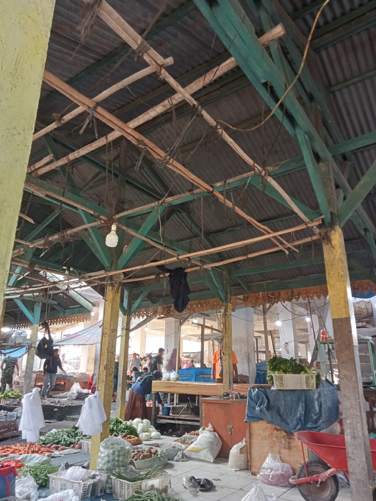 This is an image of an outdoor market in Ujung Tanjung, Indonesia. The market is covered by a corrugated metal roof supported by a wooden structure. The structure shows signs of wear and tear, with some wooden beams appearing weathered and some of the metal roofing showing rust.
In the foreground, an assortment of fresh produce is displayed on the ground and in baskets. Items include carrots, green leafy vegetables, onions, potatoes, and what appear to be sweet potatoes. Several white plastic bags and white plastic baskets are also visible, some containing produce. In the lower right of the image, a red wheelbarrow is present.
In the mid-ground, several people are visible, some appearing to be vendors and others customers. One woman in a blue headscarf and patterned skirt is interacting with produce. Another person, possibly a vendor, is standing behind a wooden table laden with more vegetables. A man in a black jacket is standing near a black bag hanging from the roof structure. In the background, more market stalls and people are visible, with glimpses of tents and other structures.
The overall scene depicts a bustling local market environment, likely a place where fresh produce is bought and sold. The lighting suggests it is daytime, with natural light filtering through the roof and the open sides of the market. The presence of hanging bags and various containers suggests active commerce.