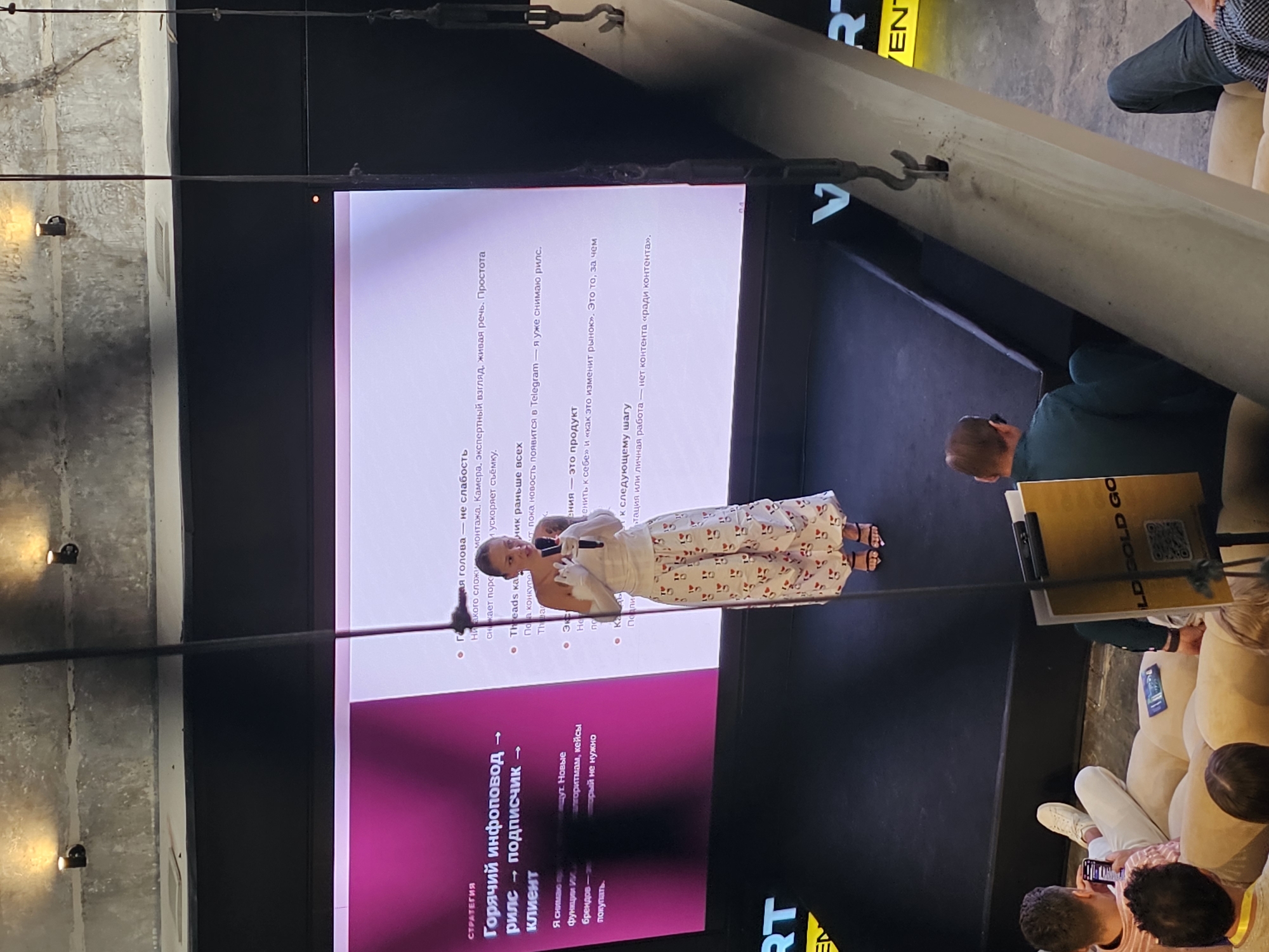 A woman is giving a presentation on stage. She is wearing a white top and pants with red hearts, and she is holding a microphone. The stage has a large screen behind her with text in Russian. The audience is seated in front of the stage. A portion of the screen shows the text "Стратегия Горячий инфоповод рилс подписчик клиент." The event is taking place indoors. The image orientation suggests the photo was taken at the city of North Kuta, Indonesia.