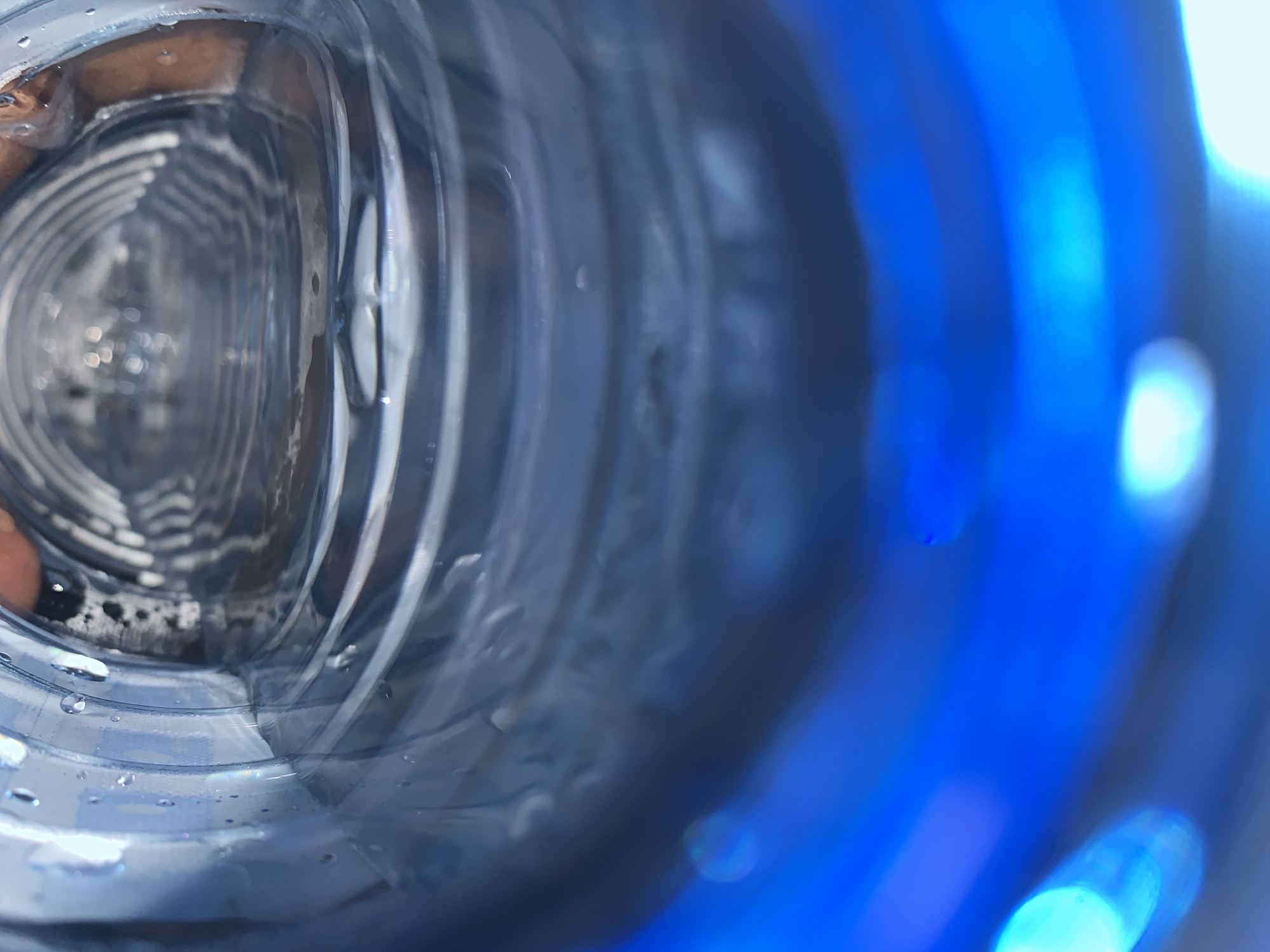 The image shows the interior of a clear plastic bottle. The view is from inside the bottle looking toward the opening. The internal structure of the bottle is visible. Droplets of liquid are adhered to the interior surface. In the foreground, partially out of focus, are sections of blue plastic. A hand holding the bottle can be seen on the left side. The location (Jos, Nigeria) is not discernible from the visual information provided.