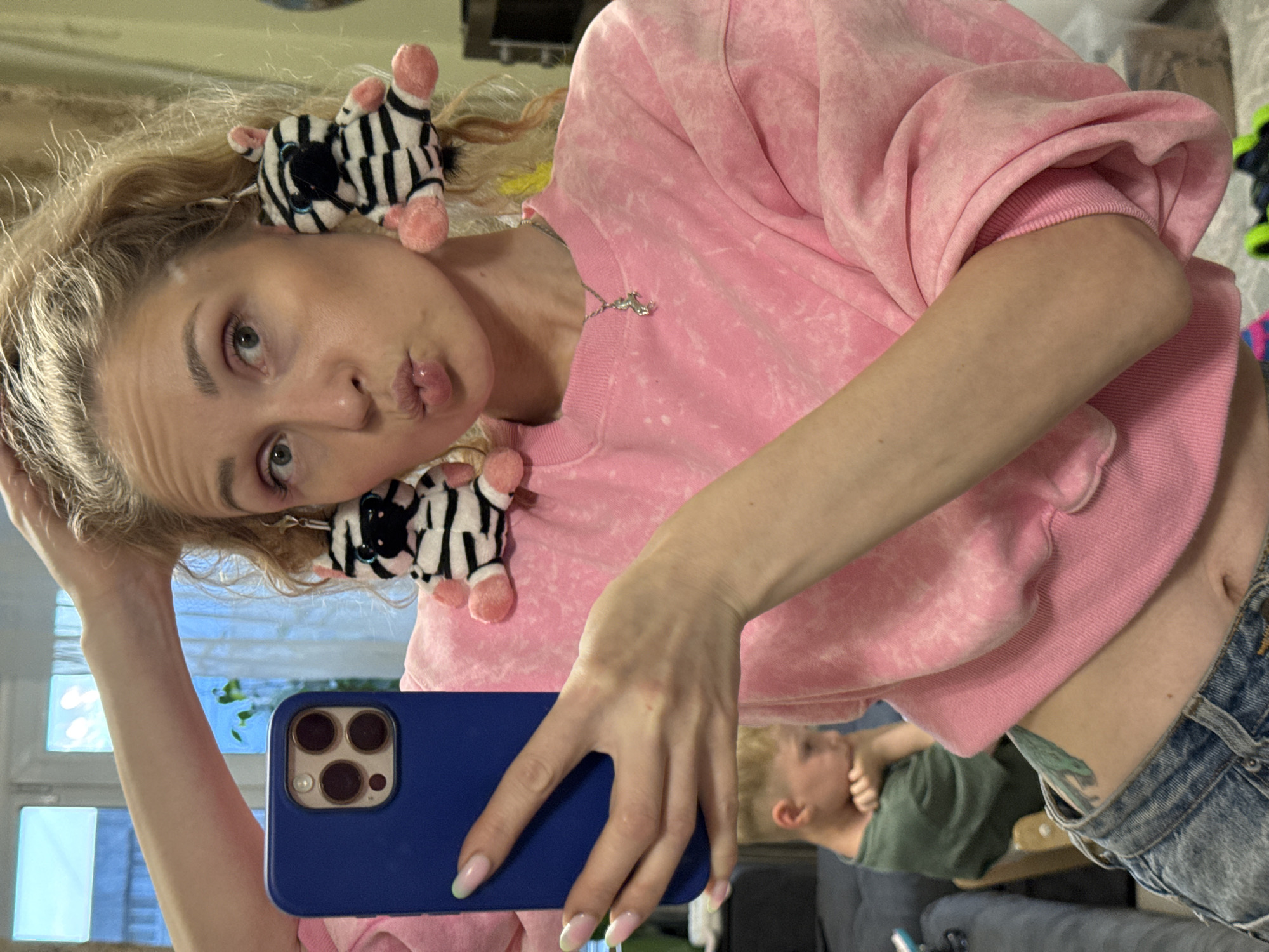 In this vertically oriented, medium shot photograph, a young woman with blonde hair styled in pigtails adorned with small stuffed zebra figures is taking a selfie in a room. She is wearing a pink tie-dye sweatshirt and blue denim shorts. Her facial expression is playful, with her lips pursed. Her left hand is holding a dark blue smartphone, angled to capture her reflection in a mirror or reflective surface.
In the background, a young boy wearing a green t-shirt is visible, also appearing to be in the same room. The room appears to be a domestic setting, possibly a bedroom or living room, with furniture and windows visible in the reflection. The lighting suggests it is daytime, but there are no specific indications of the time of day or weather conditions outside. The overall mood of the image is casual and lighthearted. No visible text is present in the image.