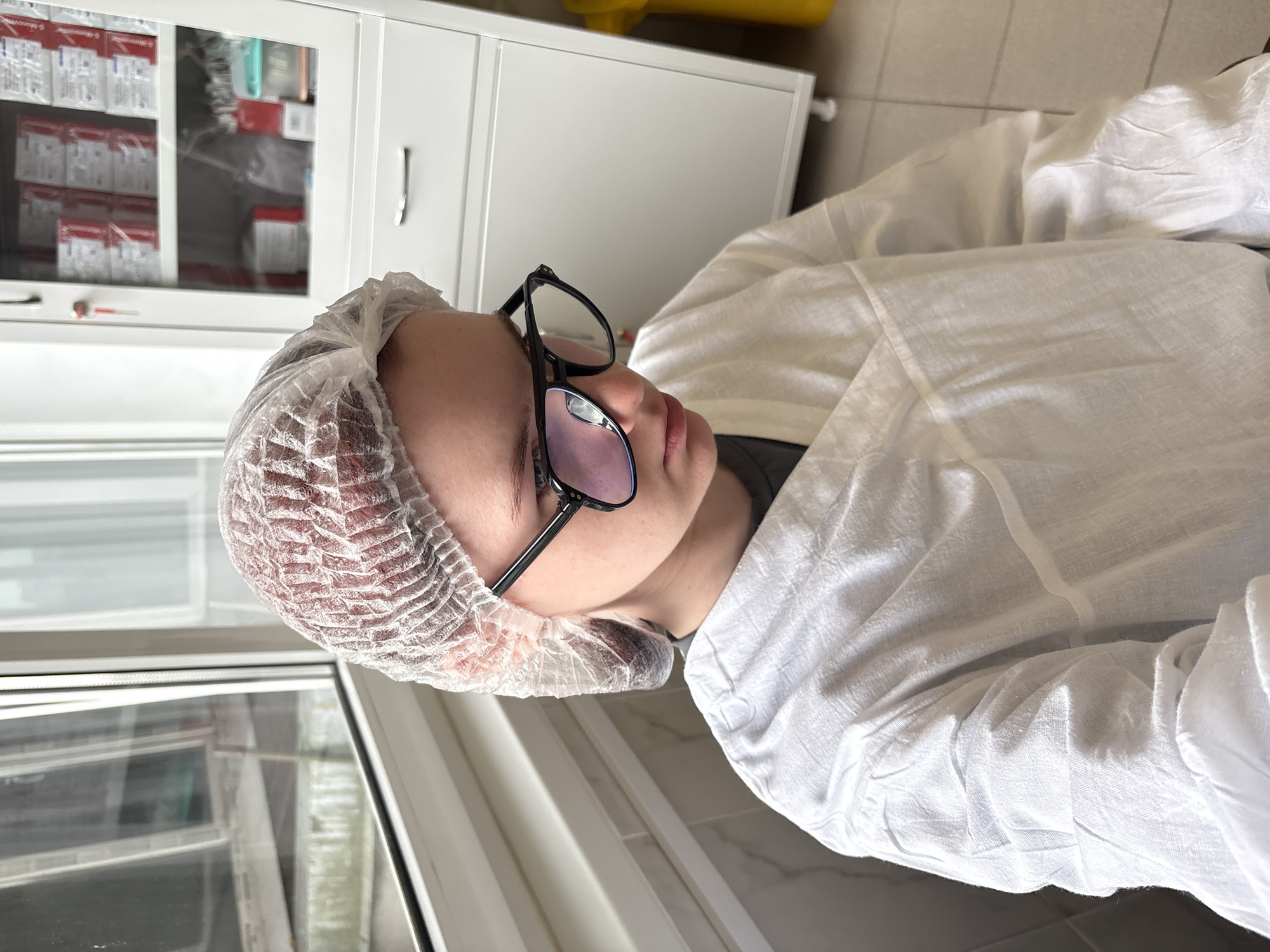 In a room with a large window and a cabinet filled with medical supplies, a person wearing a white disposable hairnet and glasses looks down. They are also wearing a white lab coat, which is partially visible. The person's head is slightly tilted to the right, and their gaze is focused downwards. The room appears to be a sterile or clinical environment, possibly a laboratory or a medical facility. The background shows a white cabinet with drawers and shelves containing various items, some in red packaging. A yellow bin is also visible to the right of the cabinet. The window on the left offers a view of an outdoor environment, though it is obscured by the reflection on the glass.