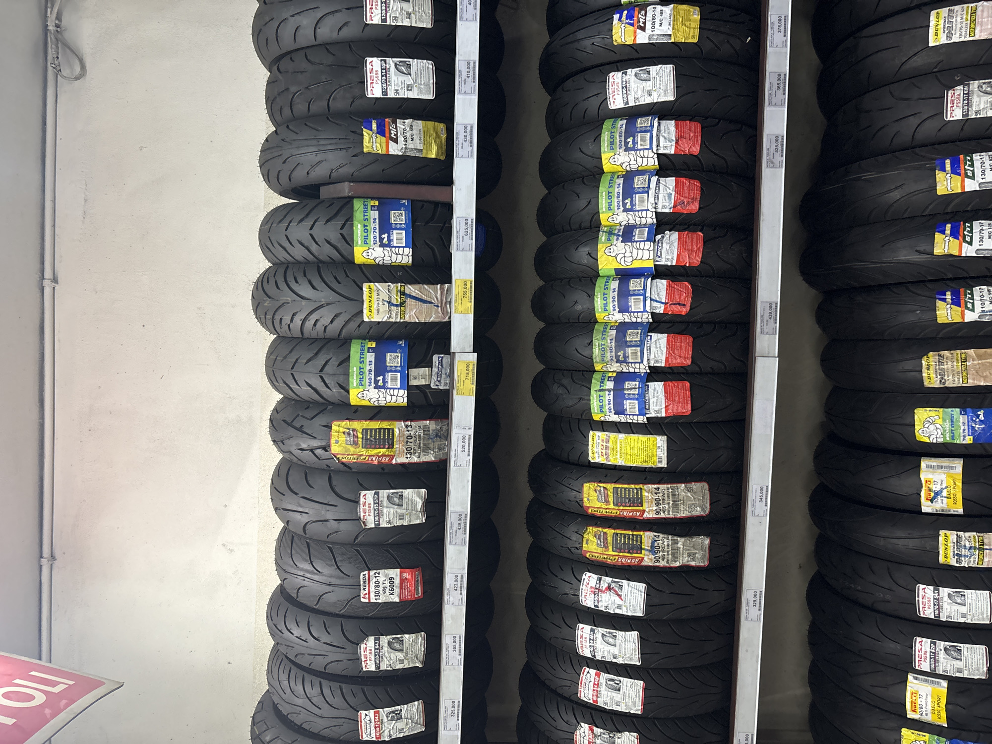 The image shows a retail display of motorcycle tires arranged on three shelves in what appears to be a store. The tires are predominantly black and feature various brand labels and specifications. The lighting suggests an indoor commercial setting, possibly a workshop or a tire retailer. The shelves are made of metal. Some tires have price tags attached. The environment seems organized and well-stocked. The image also contains partial signage in the upper left corner with the word "TOLI", which is a common abbreviation for the "Jagorawi" toll road in Indonesia, suggesting the location might be near a toll road exit or a business catering to travelers. While the specific street address is not visible, the presence of Indonesian currency denominations and labeling on the price tags suggests the location is within Indonesia, potentially in North Jakarta given the prevalence of such businesses in that area. There are no people visible in the image.