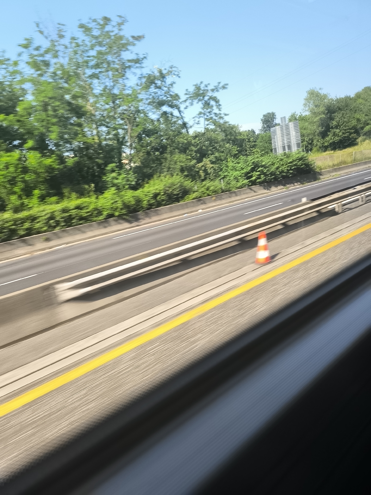The image shows a view from a moving vehicle, likely a car or bus, traveling on a highway in L'Horme, France. The highway has multiple lanes, separated by concrete barriers and a yellow shoulder line. A traffic cone is visible on the side of the road. Trees line the highway's edge. In the distance, a large, modern building with a gray, multi-story structure stands among the trees. The sky is clear and blue. The image is slightly blurred due to the motion of the vehicle.