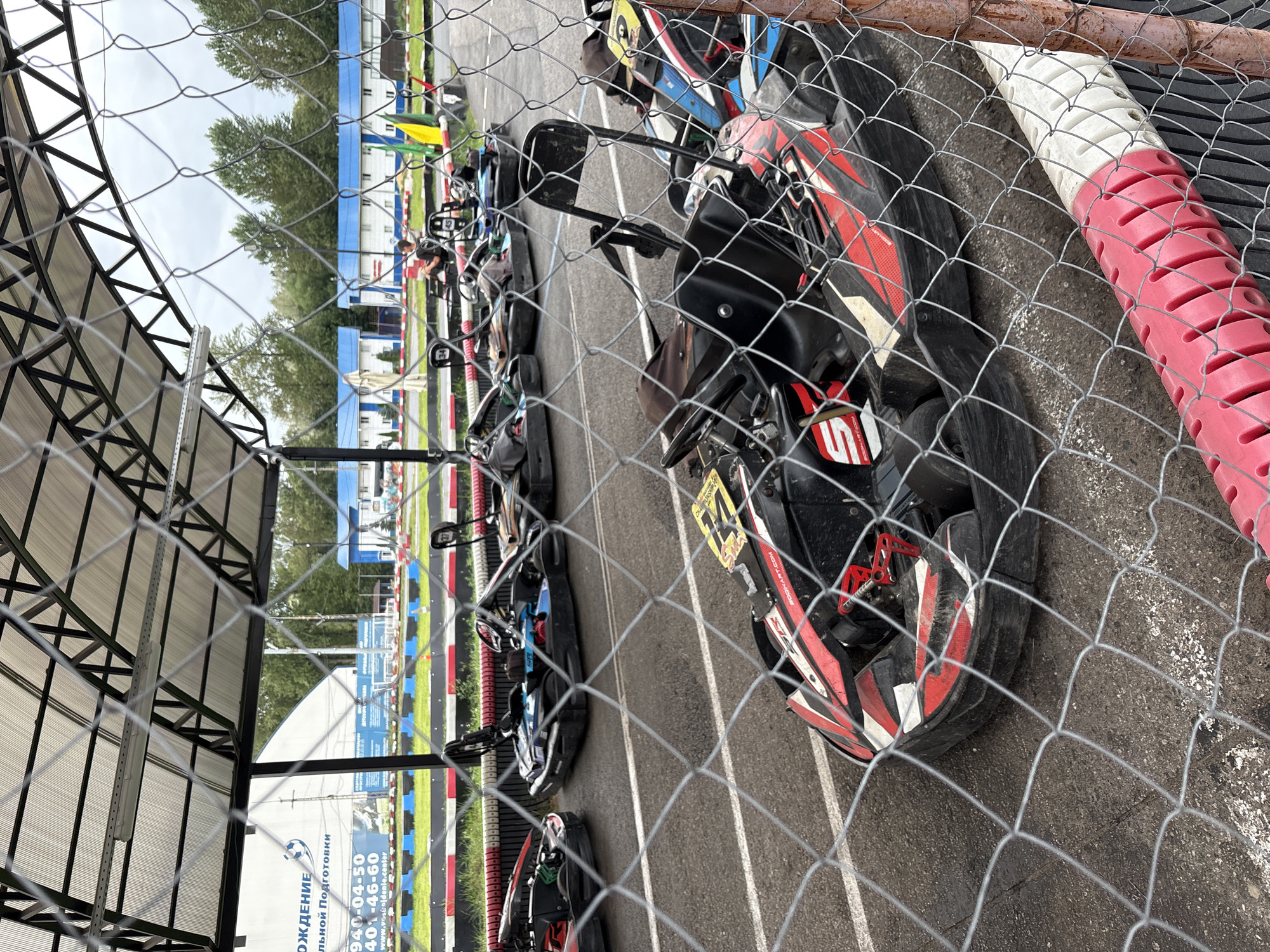 This is a photograph of a go-kart track in St. Petersburg, Russia. The image is taken from behind a chain-link fence, which partially obstructs the view of the scene. In the foreground, several go-karts are parked on the asphalt track. These go-karts are predominantly red and black, with some yellow and blue accents. They appear to be ready for use, with seats and steering wheels visible. Some of the go-karts have racing numbers on them, such as '5' and '14'. The track is lined with red and white barriers.
In the middle ground, more go-karts are visible, some of which are occupied by people. One person, wearing dark clothing, is seated in a go-kart and appears to be holding a phone or similar device. In the background, there are buildings with blue roofs, a grassy area, and trees. A large white tent-like structure with some text and graphics is also visible in the background. The sky is overcast. The overall scene suggests a recreational go-karting facility on a cloudy day.