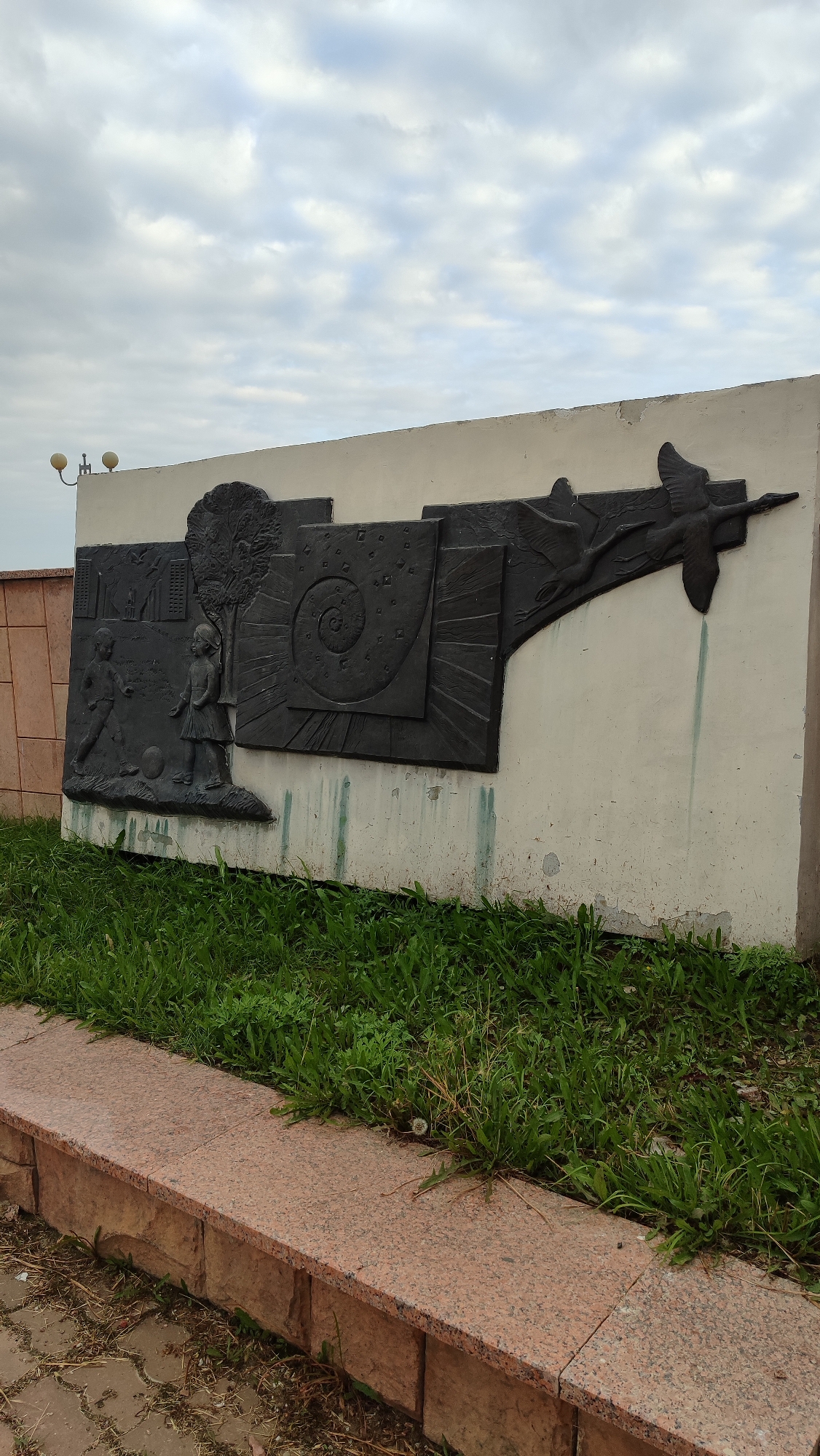 The image shows a low-angle, full shot of a bas-relief sculpture mounted on a light-colored concrete wall. The sculpture is dark gray and depicts a scene with children, a tree, geometric patterns, and flying birds. On the left, two children, a boy and a girl, are shown near a tree. The boy is kicking a ball towards the girl. Behind them, a stylized cityscape is visible. The central part of the sculpture features a spiral pattern within a square, with radiating lines. To the right, two birds in flight are depicted. The sculpture is mounted on a textured, light-colored concrete wall which appears weathered and has some green streaks running down from the sculpture. In front of the wall is a strip of green grass, with a granite border and paving stones visible in the foreground. The sky is cloudy. This sculpture is located in the city of Mirny, Russia.