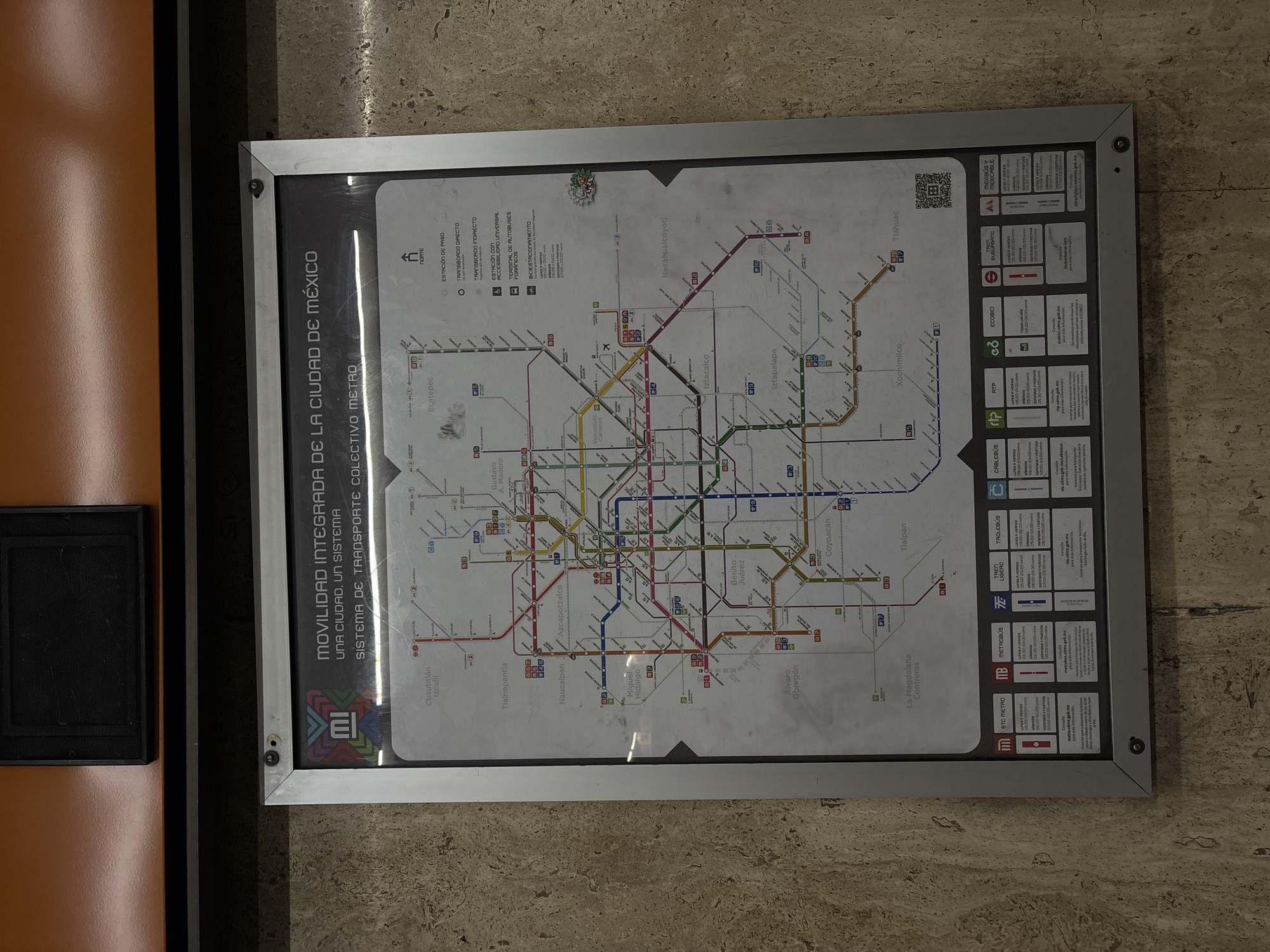 Here is a description of the image:
The image shows a framed map of the Mexico City Metro system. It is mounted on a wall, and to the left of the map is an unidentifiable black object on an orange surface. The map is mostly white with a grey border around the inside that contains the metro map itself. The map shows the various metro lines in different colors. The lines are interspersed with symbols and labels for stations, connections, transfer options, accessibility features, bus terminals, and bicycle parking. In the upper left of the map is a list of the map's features and symbols, along with information about stations, transfers, and accessibility. The bottom of the map has the logos of transportation agencies such as RTP and Metrobus, along with information about the hours of operation of the various transportation agencies in the city. The frame is silver and has screws on the corners that secure the frame in place.
