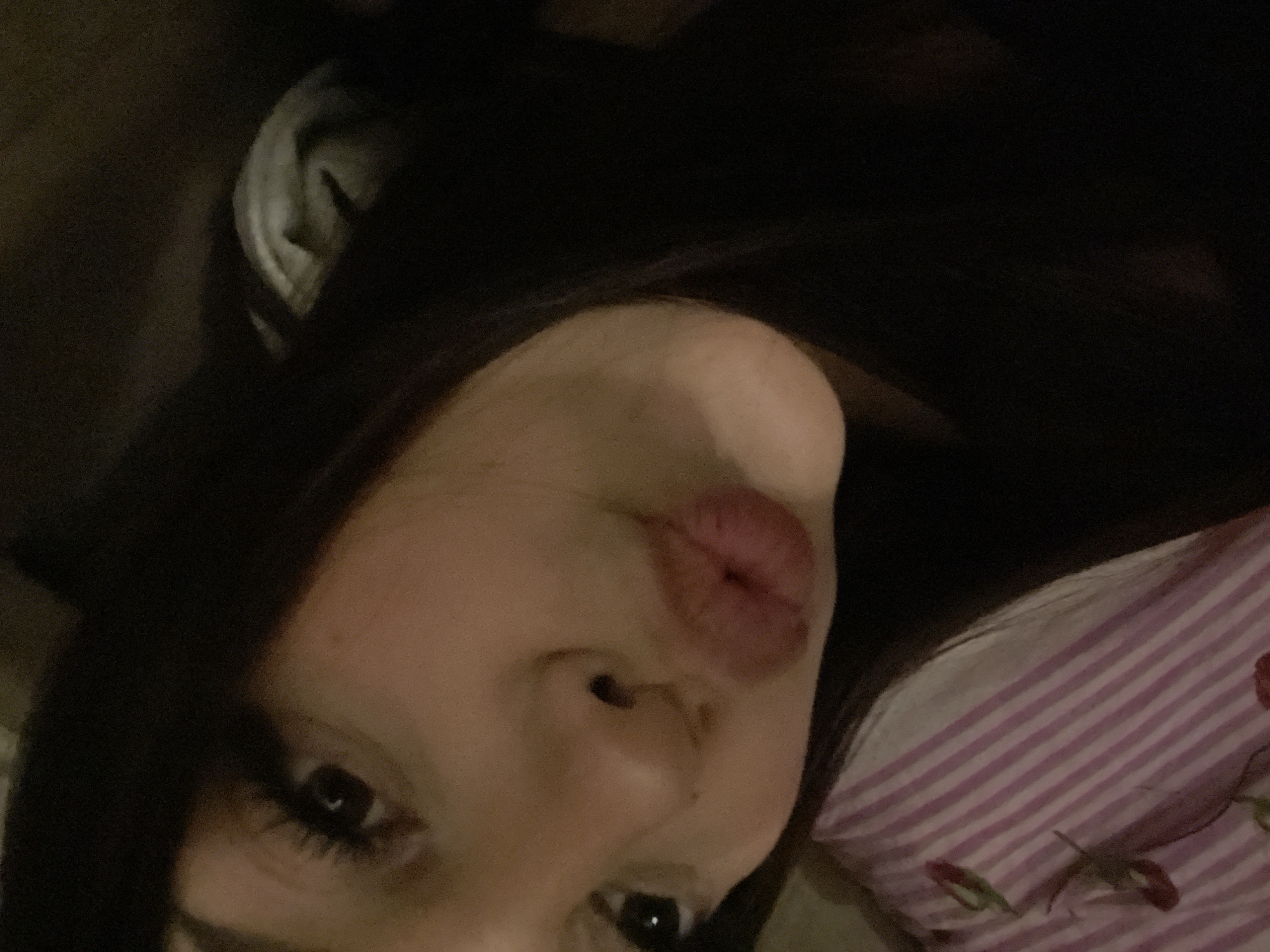 A close-up, low-light portrait captures a person, likely female, from an overhead and tilted perspective. The individual has long, dark hair that frames the left side of the face and blends into the dark background. One eye is visible, looking towards the camera, enhanced by long, dark eyelashes. The person's lips are pursed in a "kiss" or "duck face" expression, dominating the center-right of the frame. A portion of the nose is also visible below the lips.
The lighting is dim, casting soft shadows and creating a cozy, intimate atmosphere. The background is mostly dark, suggesting an indoor setting. On the right side of the image, behind the person's head, a striped fabric with light purple and white stripes is visible, adorned with red floral patterns that resemble roses. This fabric appears to be part of bedding, such as a pillowcase or blanket, indicating the person may be lying down. The overall mood is casual and playful. There is no visible text. The location context provided for this scene is Saint Petersburg, Russia.