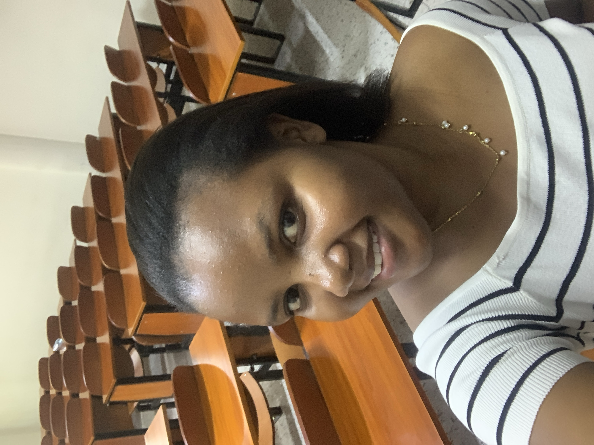 A medium shot of a woman smiling at the camera. She is wearing a white shirt with thin black horizontal stripes and a delicate gold necklace with small pearl-like embellishments. The woman has dark hair pulled back from her forehead. In the background are rows of wooden chairs typical of a classroom or lecture hall, suggesting an academic setting. The exact location within Dar es-Salaam, Tanzania, cannot be determined solely from the image, as the interior environment is not unique to the city.