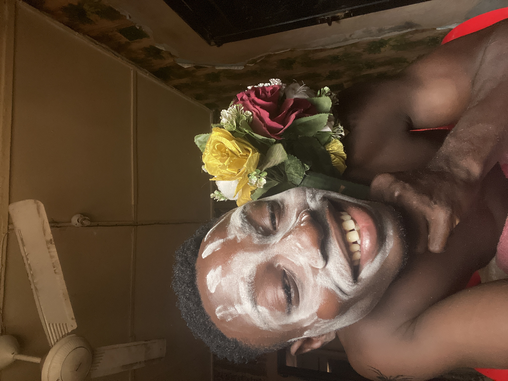 An individual with dark skin and short, dark curly hair is depicted indoors in Obosi, Nigeria. The person is applying a white substance, likely a face mask, in streaks across their forehead, cheeks, and chin. They are smiling broadly, revealing their teeth, and their eyes are closed or squinting in apparent joy or amusement. They are holding a bouquet of artificial flowers, prominently featuring red and yellow roses along with smaller white blossoms and green foliage, positioned near their head.
The setting appears to be a rustic indoor space. The background features light brown or beige walls, possibly with vertical paneling. The ceiling, visible on the right side of the frame due to the image's orientation, shows an uneven, textured surface with dark, discolored patches. A dusty, white ceiling fan with two blades is visible on the left, mounted near the wall, along with a small, white light fixture above it. Part of the person's bare shoulder and arm are visible on the right, suggesting minimal clothing, with a hint of red fabric. A faint tattoo-like marking is visible on their arm. The overall atmosphere conveys a sense of playfulness and self-care. There is no visible text in the image.