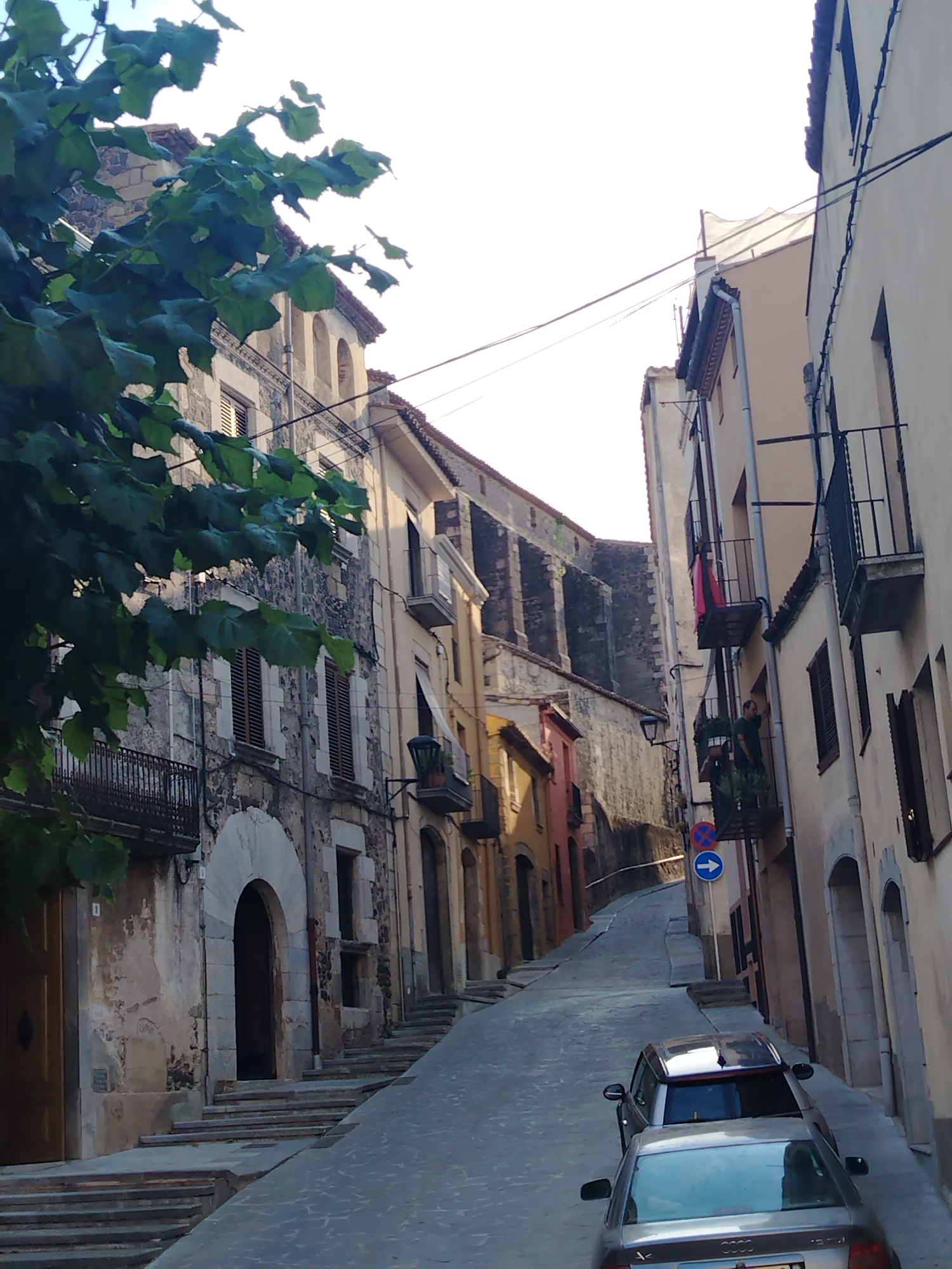 The image depicts a narrow, sloping street in the historic town of Hostalric, Spain. The street is paved with cobblestones and lined with old buildings made of stone and plaster, displaying signs of age with peeling paint and weathered facades. On the left, a building features an arched entryway and stone detailing. Green foliage from a tree hangs into the frame from the upper left. Two cars are parked on the street, one silver sedan in the foreground and a darker SUV behind it. The buildings on either side have balconies with wrought-iron railings, and several windows are visible, some with closed shutters. A man is seen on a balcony on the right side of the street, looking out. Traffic signs are posted along the street. The sky above is bright and appears to be a clear day, with some overhead wires present.