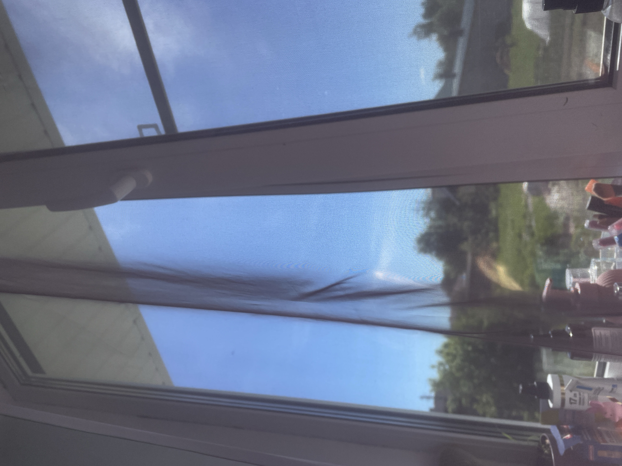 The image displays an outdoor view seen through a wide, horizontally oriented window, which is covered by a fine mesh screen. A white window frame or mullion horizontally divides the image.
Through the screen, a bright blue sky with faint white clouds is visible in both the upper and lower sections of the window. Below the sky, in the lower part of the view, green foliage, including trees and grass, suggests a garden or outdoor area. The light-colored roof of a building is faintly visible in the top-left corner through the screen.
The white window frame is clean and features a white handle on its upper-left section, indicating the window is operable. The mesh screen covering the entire window gives the outdoor scene a textured, diffused appearance, and it shows a noticeable sag or crinkling in its central portion.
To the far right, beyond the window frame, several indistinct objects are visible. These appear to be bottles or containers of various shapes, likely placed on a windowsill or shelf indoors. No legible text is discernible on any of these items.
The clear blue sky and bright lighting indicate a sunny daytime setting. The scene provides a glimpse of a garden or suburban environment, located in Медведево, Russia.