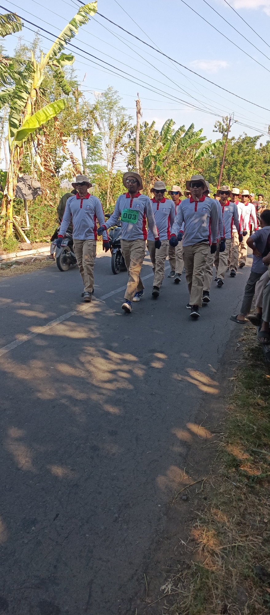 In Ketowan, Indonesia, a group of men are participating in a walking event. They are dressed in matching light gray long-sleeved shirts with red collars and accents, tan cargo pants, hats, and dark gloves. Some of them are wearing sunglasses. One man in the front row has a green bib with the number "003" on it. They are walking in a line down a paved road lined with trees and vegetation. In the background, a motorcycle is parked on the side of the road. The sun is shining, casting shadows on the road. There are also spectators on the right side of the road, some of whom are also dressed in similar attire.