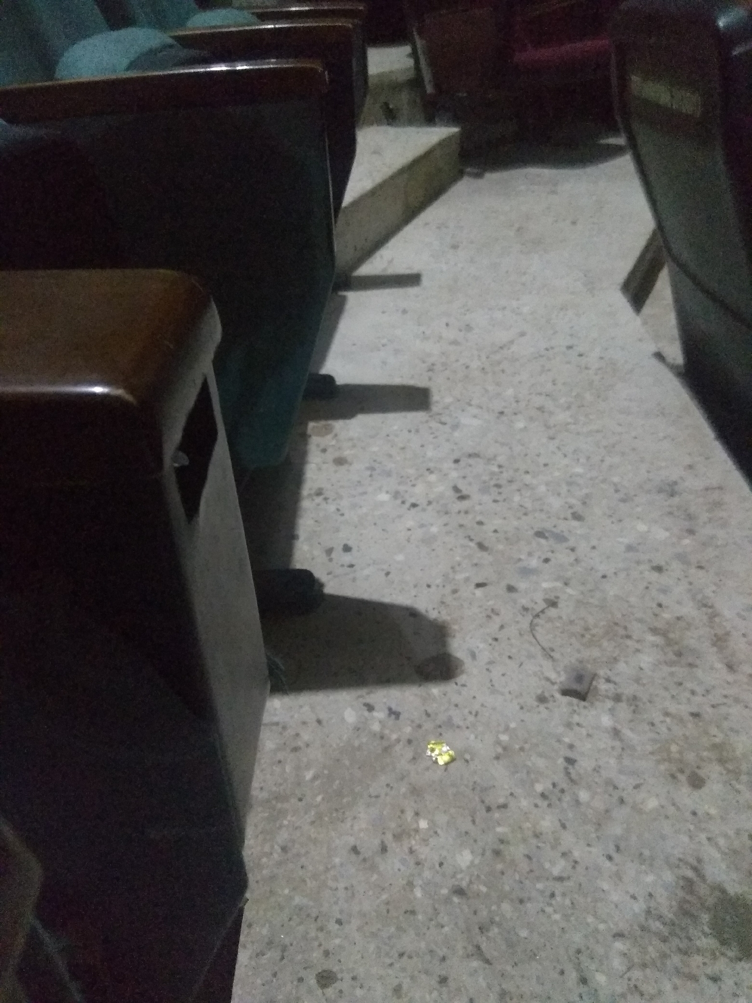 The image depicts an interior scene, likely a theater or lecture hall, with rows of empty seats and a textured floor. In the foreground, a portion of a dark-colored seat armrest is visible, with a small, reflective object on the floor near it. The floor is a light gray concrete with dark specks, typical of public spaces. Shadows from the seats are cast across the floor, suggesting the presence of overhead lighting, though none is directly visible. The background shows more seats and a raised area that might be a step or stage. The overall impression is one of quiet and disuse. The context of Jalingo, Nigeria, would place this scene in a specific urban environment within that country, potentially within an educational institution, entertainment venue, or government building.