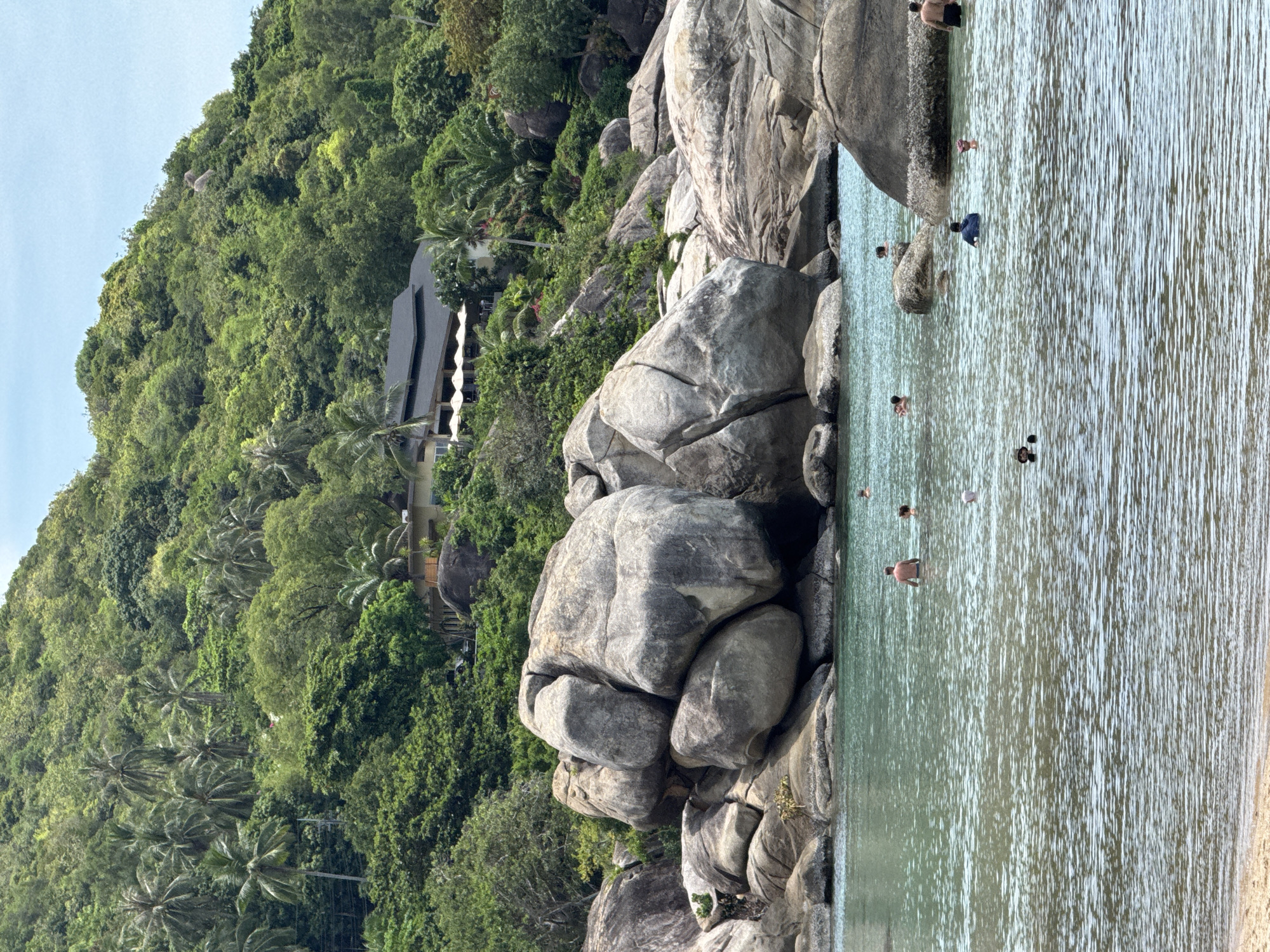 The image depicts a scene at Baan Chaweng Noi, Thailand. In the foreground, there is a body of water, likely the sea, with several people swimming or wading. Large rock formations are present along the shoreline, creating a boundary between the water and the land. Lush green vegetation covers a hillside in the background. A multi-story building is visible amongst the trees. The scene is bathed in natural daylight, indicating a sunny or bright day.