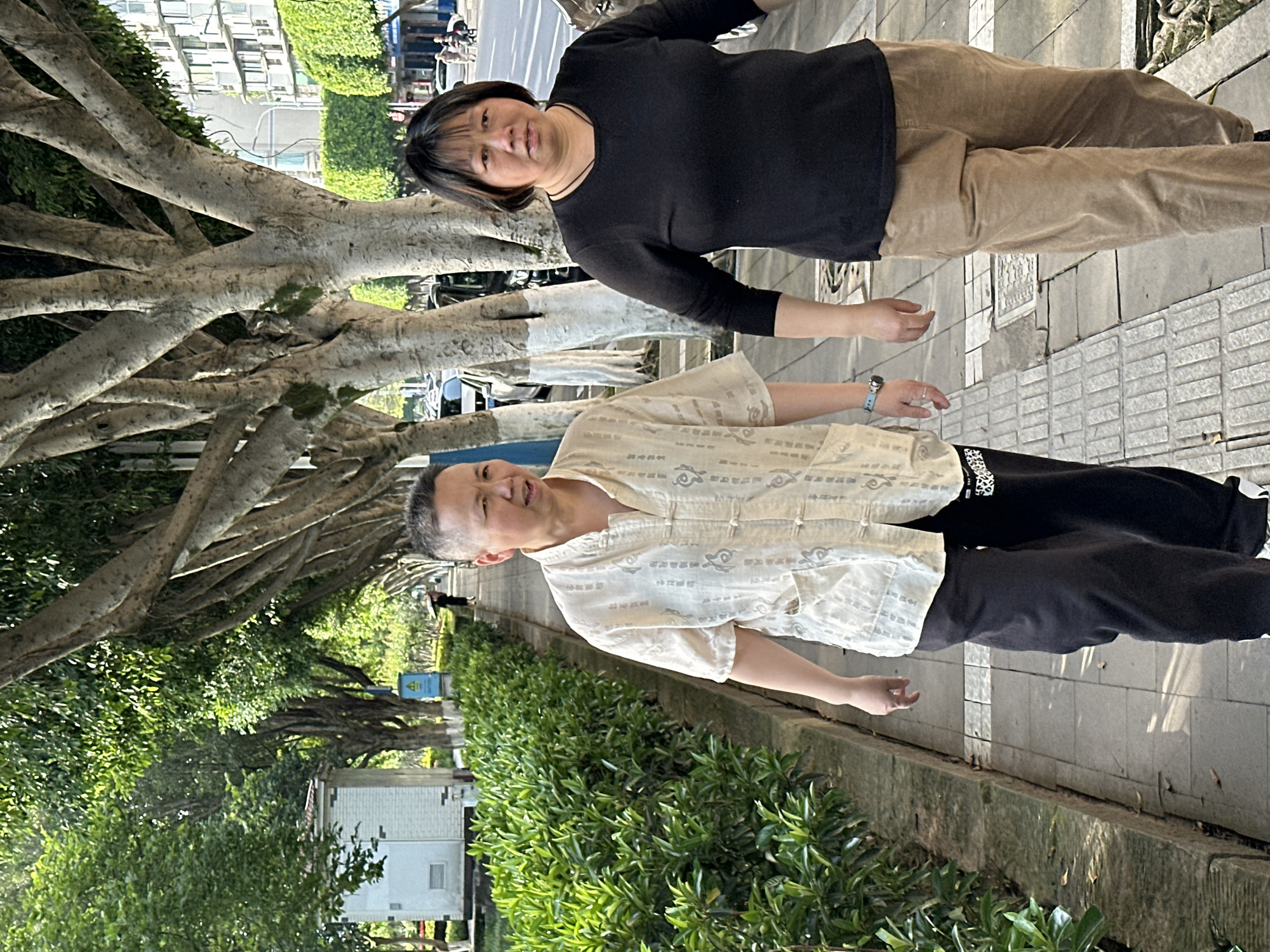 Two people stand on a paved sidewalk in Jiangjin, China. To the left, a large tree with exposed roots and branches extends over the walkway. A woman in a black long-sleeved shirt and khaki pants stands on the right of the frame. Next to her is a man in a white patterned shirt and black pants. A concrete curb separates the sidewalk from a row of green bushes. Further back, a white building is visible. A street runs parallel to the sidewalk with several parked cars. Additional buildings are visible in the background.