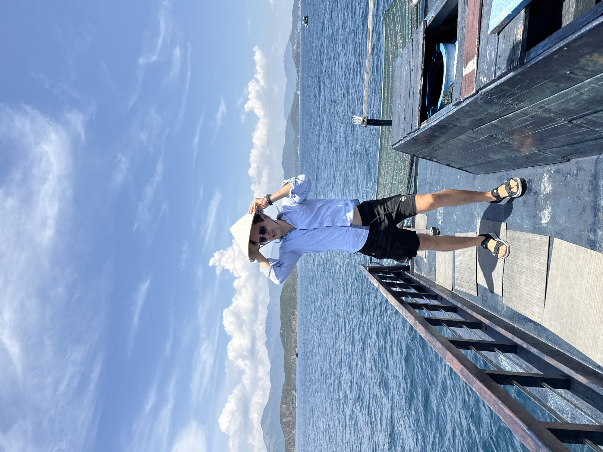 The image shows a man standing on the side of a boat. He is wearing a light blue button-down shirt, black shorts, sandals, sunglasses, and a conical Asian hat. He is holding the hat with both hands on top of his head. The boat is docked or anchored in the ocean, and the background shows a clear blue sky with fluffy white clouds, a body of water, and distant mountains. Part of a ladder is visible, extending from the boat towards the water. The location is Nha Trang Ward, Vietnam.