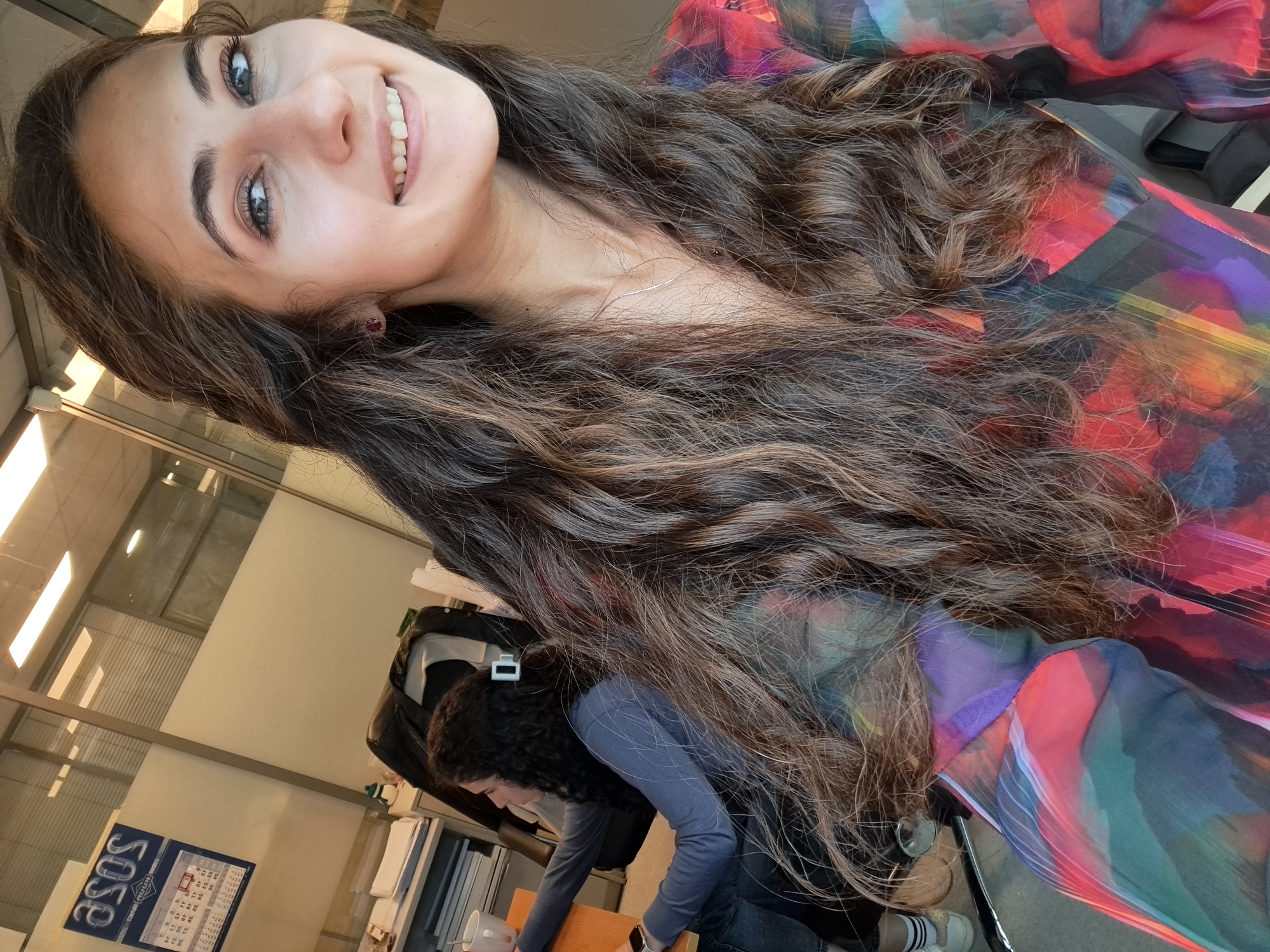 The media file displays an indoor scene, likely an office or modern workspace in Sofia, Bulgaria, characterized by bright lighting and glass partitions. The image is rotated 90 degrees counter-clockwise, making the main subject appear to be looking upwards or sideways from a lower position.
The primary subject is a young woman with long, wavy brown hair, featuring lighter highlights. She is smiling broadly at the camera, revealing her teeth, and has expressive blue or green eyes and dark eyebrows. She wears a vibrant, sheer blouse patterned with red, pink, purple, blue, and green hues. A delicate silver necklace is visible around her neck, and she has a small red stud earring in her left ear. Her head is tilted, giving a dynamic feel to the portrait.
Partially visible in the background is a second person with dark curly hair, wearing a long-sleeved blue-grey shirt and dark pants. This individual appears to be leaning forward, possibly working at a desk or table, with a white hair clip visible in their hair. Behind this second person, elements like a black backpack, a white mug, and stacks of documents or binders on shelves can be seen. The floor is light-colored, and a pair of white sneakers with black-striped socks is visible near the second person's feet.
Notable details include the bright, daylight-like ambiance, suggesting it is daytime. The main subject's cheerful expression conveys a positive emotion. On a blue sign or poster in the background, the word "ASOS" is clearly visible, along with a grid that resembles a calendar.
