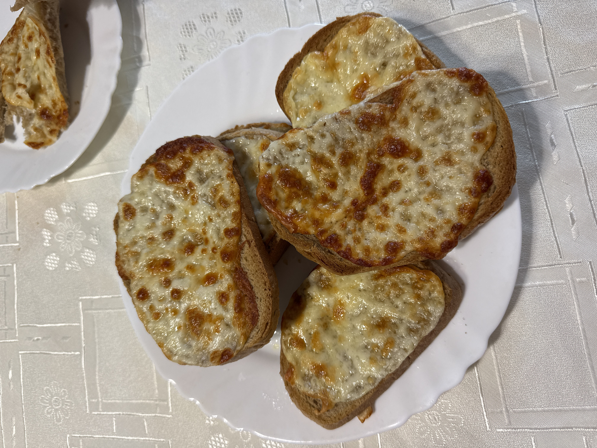 The image shows a white plate holding five pieces of toasted bread topped with melted cheese. The bread is golden-brown with a slightly charred appearance around the edges. The cheese has a bubbly texture with brown spots. A second plate with similar toast can be seen in the upper-left corner of the image. The plates sit on a table covered with a white, patterned tablecloth. The scene does not contain any information about the location of Blagoevgrad, Bulgaria.