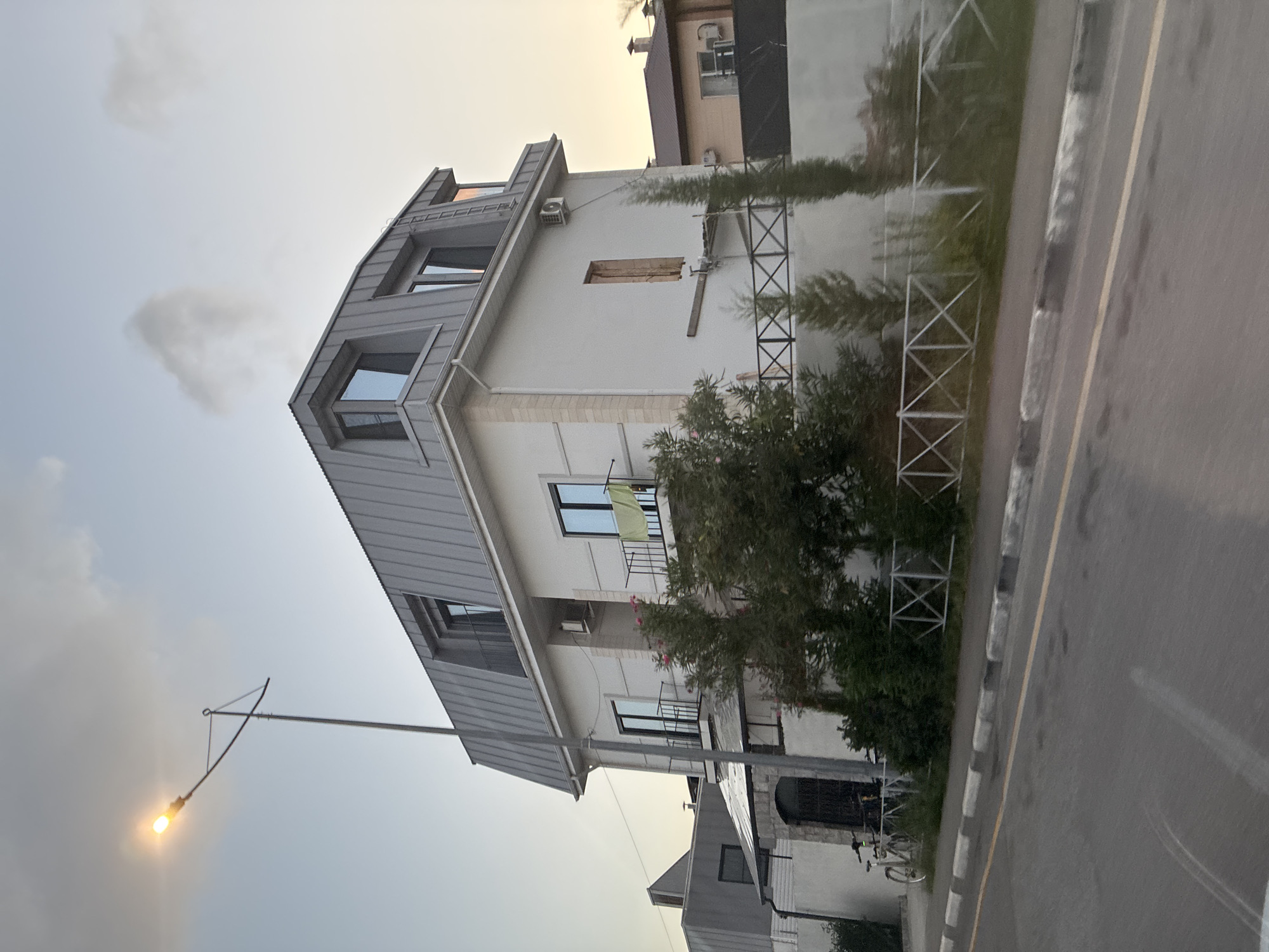 A street-facing, multi-story white building with a mansard roof is visible. The building has numerous windows, some with balconies. A street light with a lit bulb is positioned to the left of the building. Adjacent to the building is a paved road with a curb. In front of the building, there is a metal fence enclosing a garden with trees and bushes. A bicycle is parked near the entrance of the building. The sky is overcast with a few clouds, and the lighting suggests it is either early morning or late evening. This scene is located in Sirius, Russia.