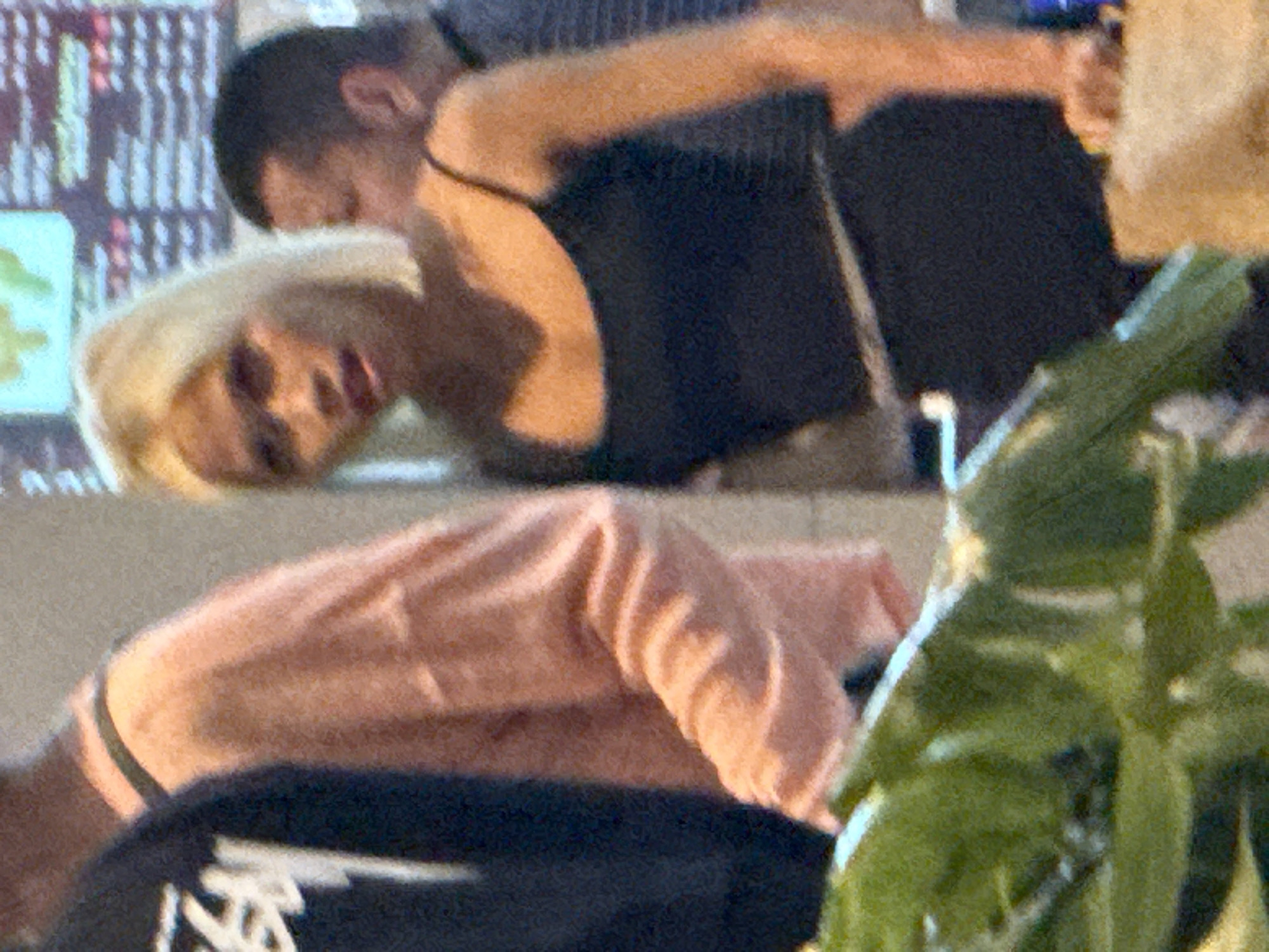 The image displays a group of individuals indoors, likely in a casual dining establishment given the blurry background signage that resembles a menu. In the foreground, a woman with short, blonde hair and fair skin is centrally positioned, wearing a black tank top and black bottoms. Her gaze is directed downwards, and her mouth is slightly open. To her left, partially visible, is another person in a light pink, long-sleeved shirt. To the right of the blonde woman, a person with short, dark hair and fair skin is visible from the side, wearing a dark garment. In the far left of the image, a person in a black t-shirt with a white graphic is partially visible. The environment appears to be a public indoor space, possibly a restaurant or food court, with some foliage in the lower left corner. The image quality is grainy, obscuring fine details. There is no clear indication of the location being specifically in Bắc Cam Ranh, Vietnam.