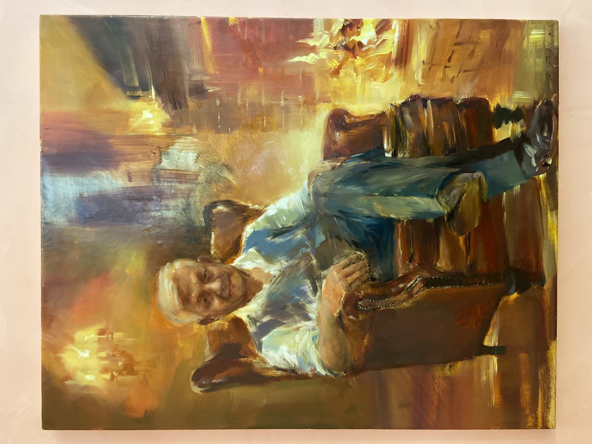 A painting depicts a man reclining in a brown leather chair, with his legs crossed and resting on a stack of books or boxes. The man is wearing blue jeans and a light-colored shirt. The background is rendered in warm tones of yellow, orange, and brown, creating a soft and atmospheric effect. The overall style appears impressionistic, with visible brushstrokes. The painting is signed "A. Zak. 2018" and is potentially located in Kazan, Russia.