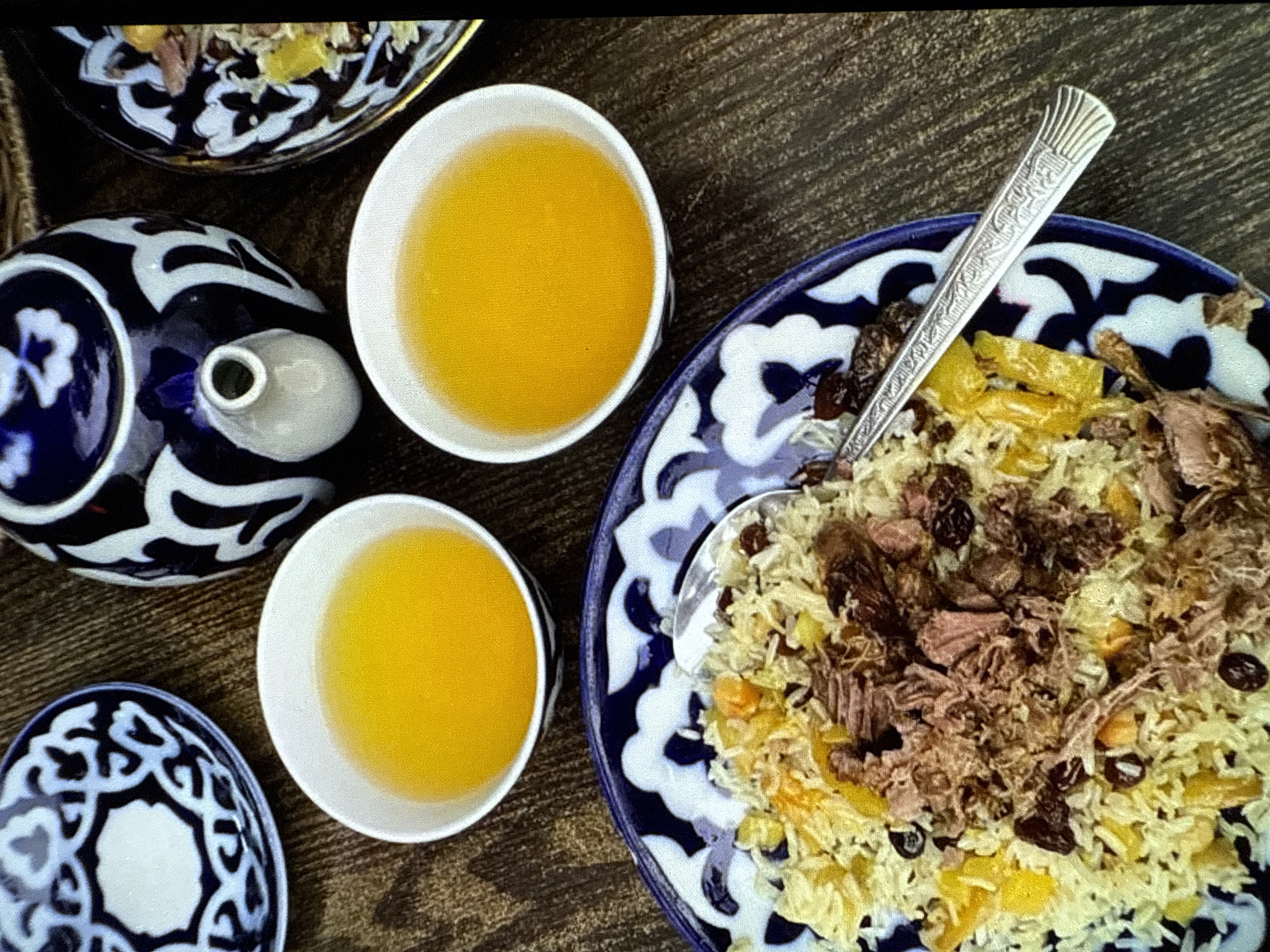 An overhead shot shows a meal laid out on a wooden table. In the foreground is a blue and white patterned plate piled high with pilaf, a rice dish mixed with meat, raisins, and yellow pieces, possibly carrots or bell peppers. A spoon rests on the edge of the plate. To the left and right of the pilaf plate are two white bowls filled with a golden liquid, likely tea. Behind the bowls and pilaf plate is a white and blue patterned teapot. To the far left and right are two more blue and white patterned plates, one of which contains pilaf. The overall setting appears to be a traditional or cultural dining experience.