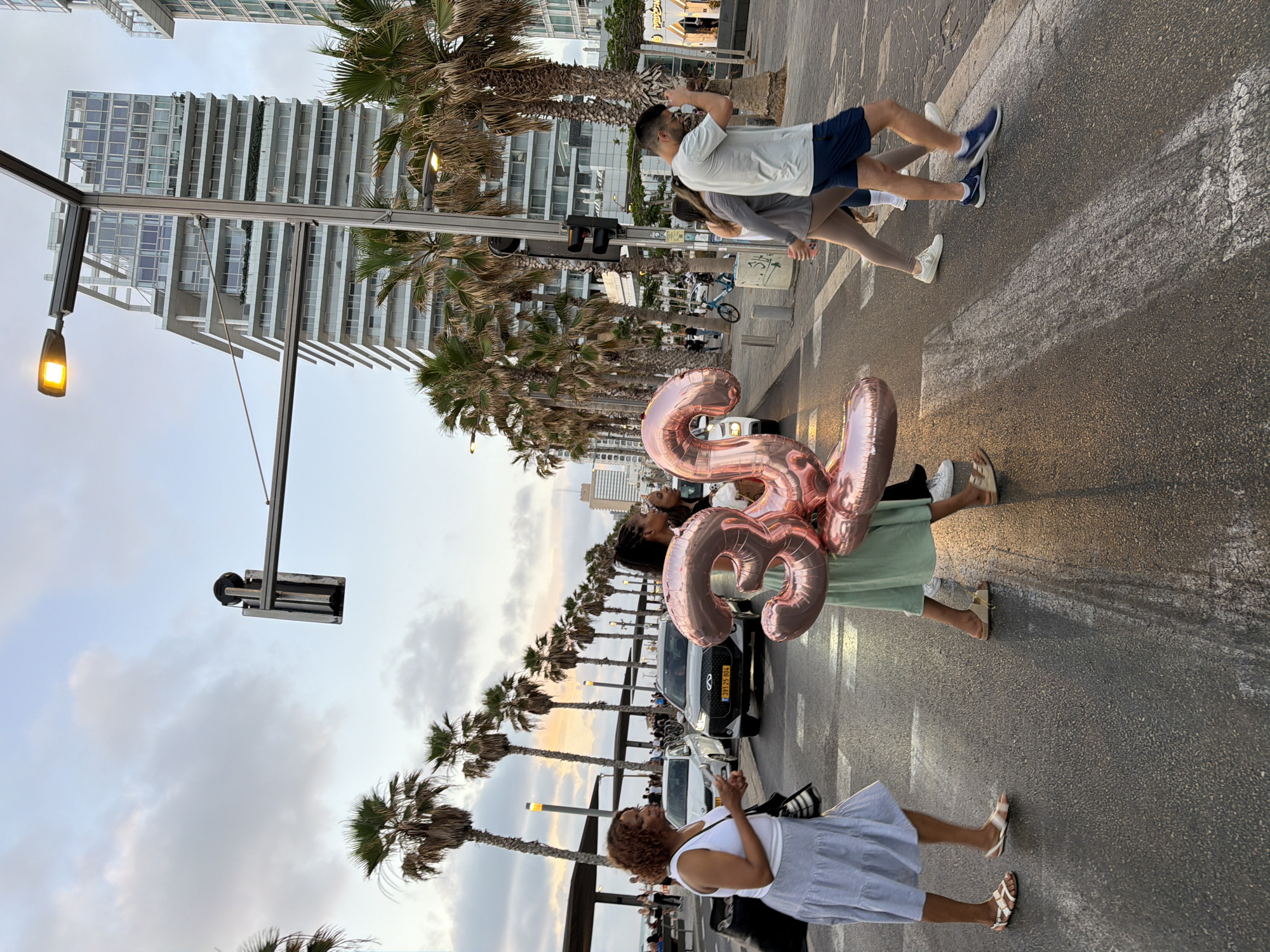 The image depicts a street scene likely in Tel Aviv, Israel, given the architecture and Hebrew license plates on a vehicle. The setting is a wide sidewalk or promenade alongside a palm tree-lined street. Several people are present: one woman holds large, pink, foil "33" balloons, possibly celebrating a birthday; another woman in a white top and blue skirt stands nearby; and in the background, two people are crossing the street. The building's architectural style and the presence of palm trees suggest a coastal, urban environment typical of Tel Aviv. Streetlights and traffic signals are also visible. The overall atmosphere appears casual and celebratory.