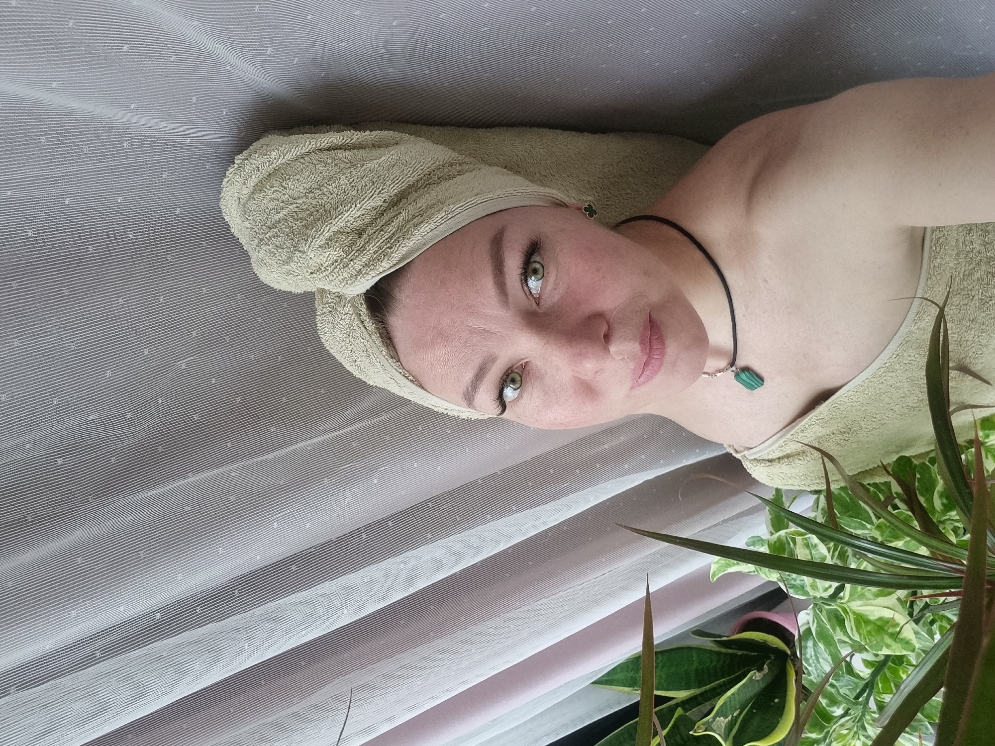 A woman with a towel wrapped around her head and a towel draped over her shoulders is looking into the camera. She has light skin, green eyes, and pink lips. She is wearing a black cord necklace with a green pendant. A houseplant with long, pointed leaves is in the lower right corner of the image. A sheer, white curtain hangs in the background. The location of the image is not provided.