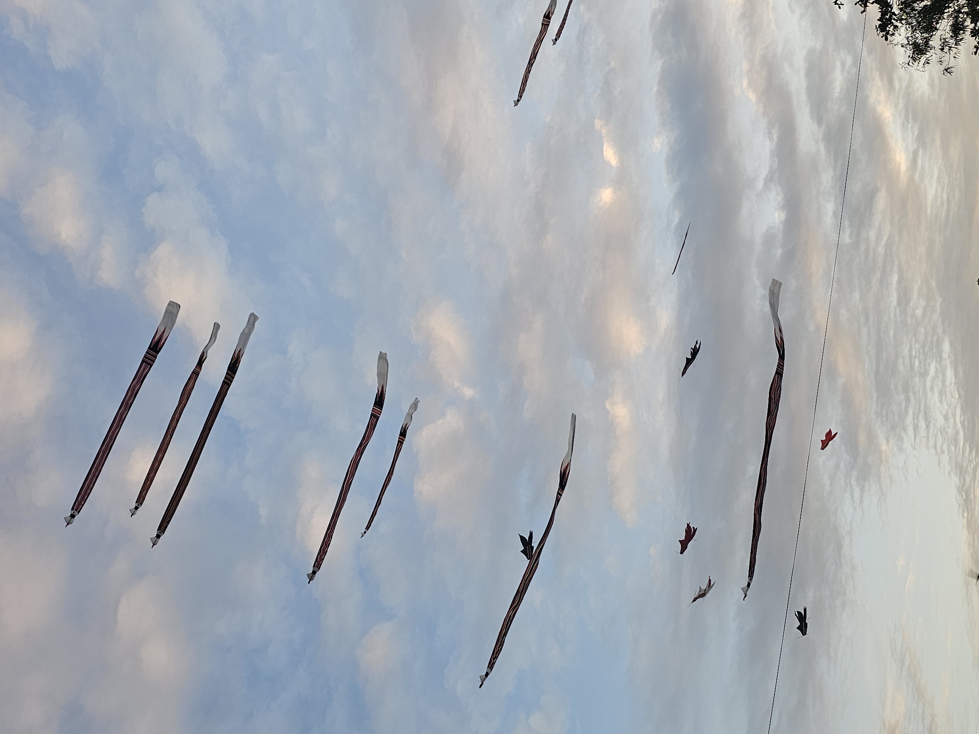 The scene is a low-angle shot of the sky in Denpasar, Indonesia. The sky is filled with white and light blue clouds. Several long, narrow, striped kites or banners are flying in the sky. Some smaller, more detailed paper figures that resemble birds or bats are also attached to or flying near these banners. The scene is most likely an outdoor festival.
