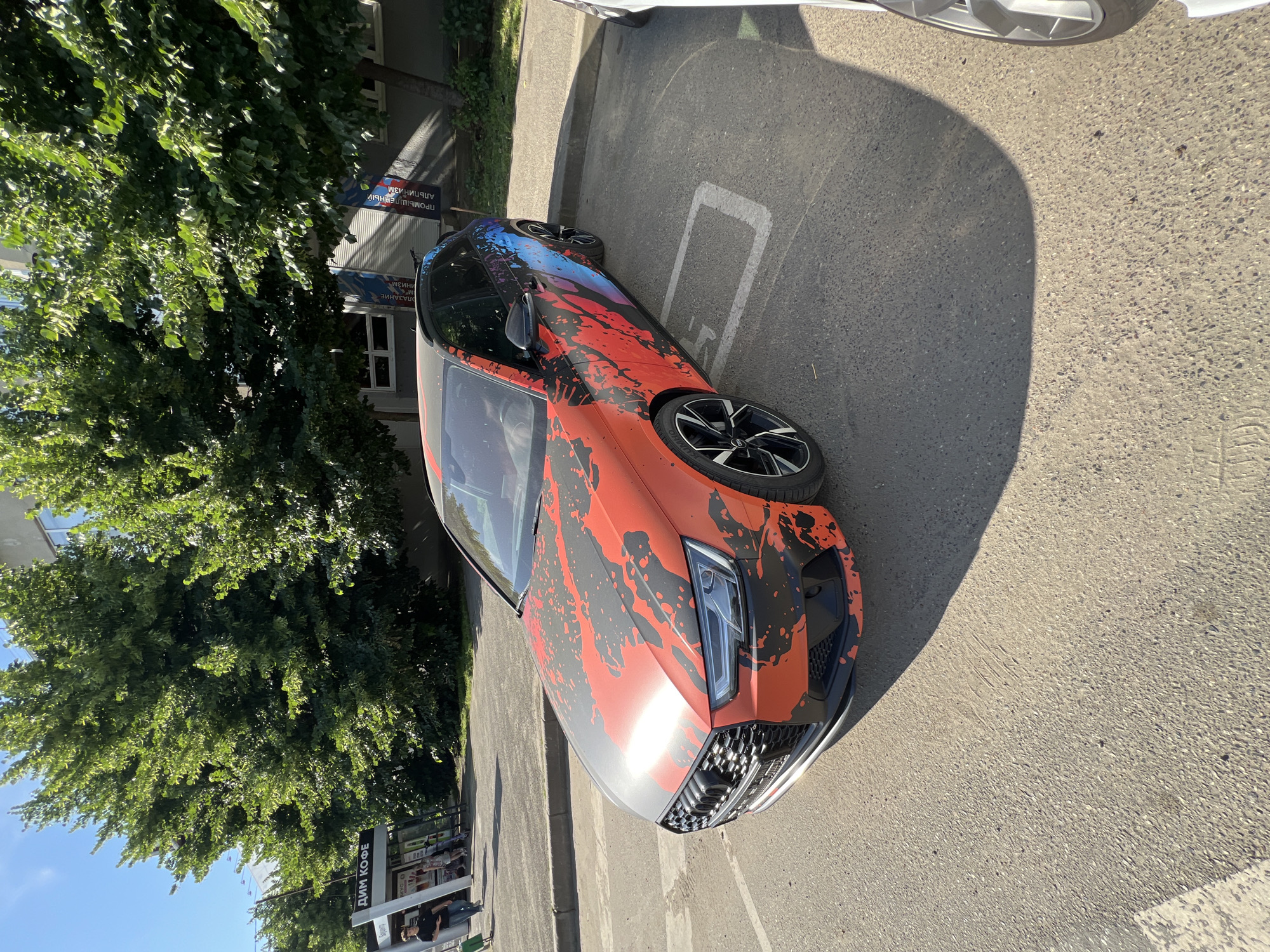 An orange and black Audi A4 Avant is parked in a lot outside a building in Krasnodar, Russia. The car has a custom wrap with a red and black splatter design on the hood and sides, transitioning to a blue and purple abstract design on the rear. The car is parked in a designated parking spot, indicated by white lines on the asphalt. In the background, there are trees and a building with windows. To the left of the car, there is a bus stop or kiosk with signage in Russian. A few people are visible walking near the kiosk. The weather appears sunny, casting a shadow from the car onto the ground.