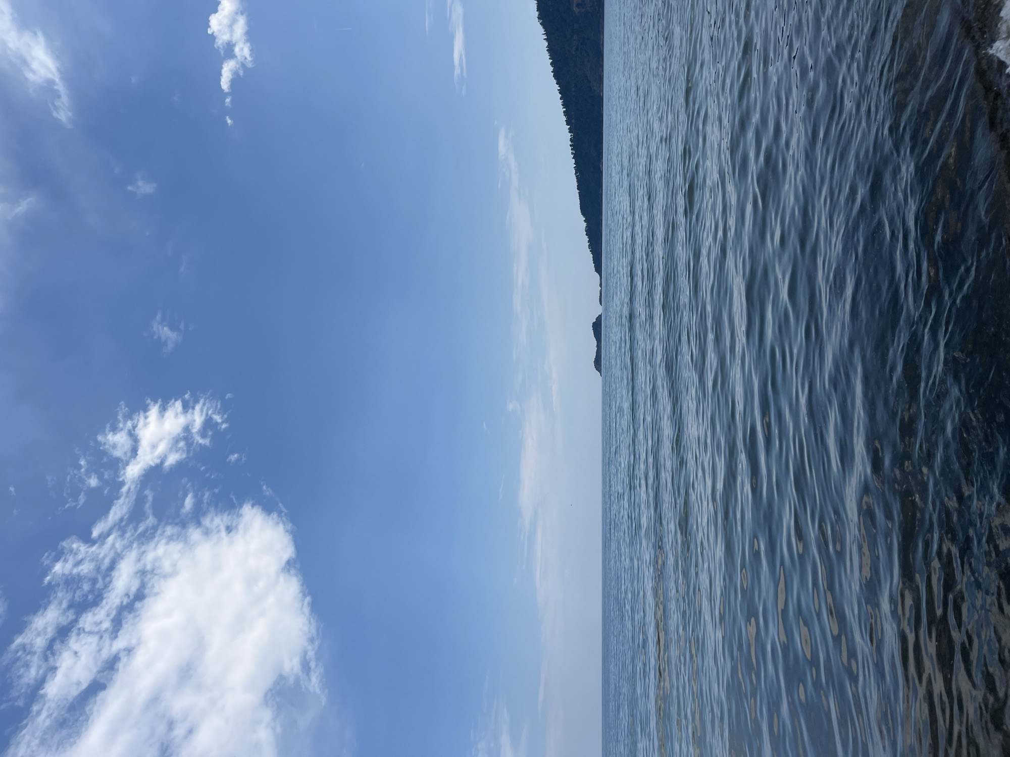 The image depicts a serene seascape with a clear blue sky filled with scattered white clouds. The foreground is dominated by gentle waves lapping at a pebbly shore. In the distance, a tree-covered coastline stretches across the frame, with a small, rocky islet visible just offshore. The horizon line is crisp, separating the calm water from the expansive sky. The scene suggests a peaceful day at the beach in Golden Beach, Greece, a location known for its natural beauty and tranquil atmosphere. There are no people or animals visible in the image. The overall impression is one of natural beauty and tranquility.