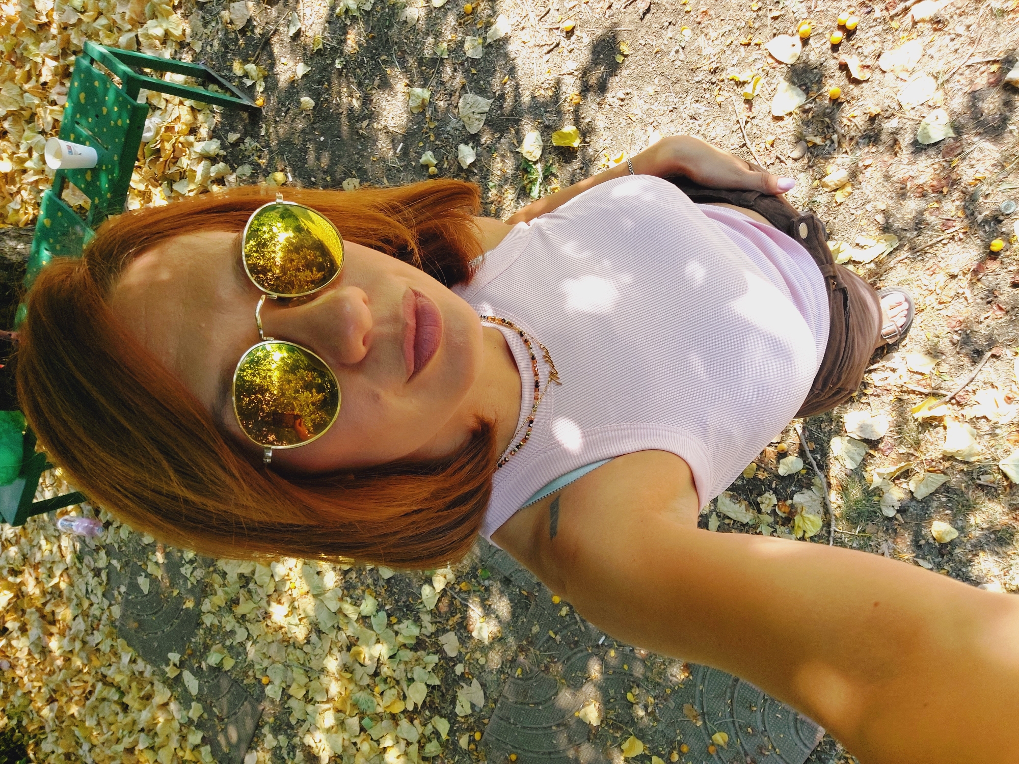 A woman with auburn hair, wearing round, gold-rimmed sunglasses, a light pink ribbed tank top, and brown shorts, is taking a selfie outdoors. She is standing on a path covered with fallen leaves and small yellow fruits. In the background, a green park bench with a white cup on it is visible. The scene appears to be in a park in Sofia, Bulgaria, suggested by the presence of ginkgo leaves and the specific type of park bench common in Bulgarian cities. The sunlight casts dappled shadows on the ground.