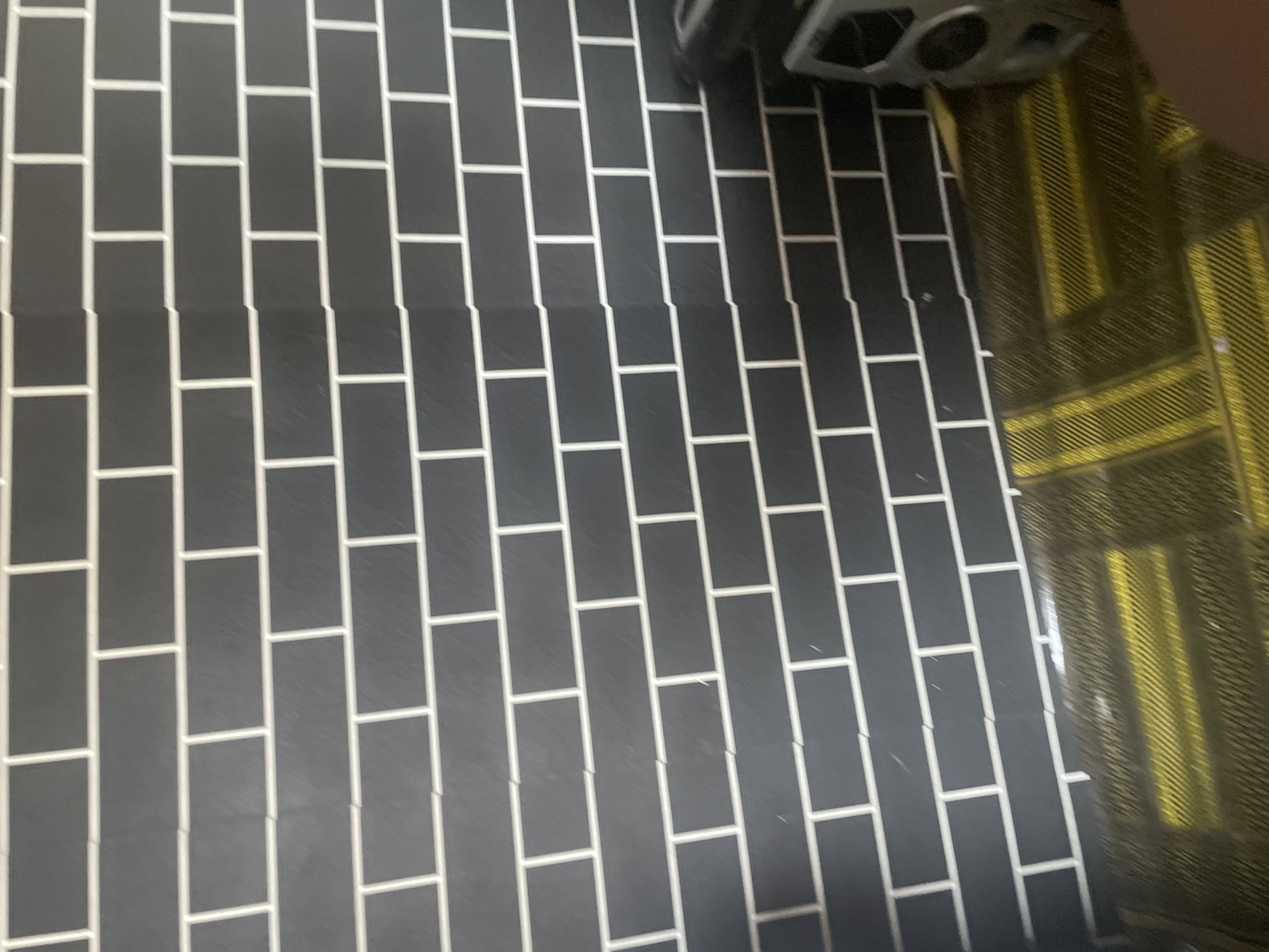 The image shows a close-up view of a floor covered in dark rectangular tiles with white grout lines, arranged in a staggered brick pattern. The tiles occupy the majority of the frame, with a portion of a yellow and black patterned rug visible in the upper right corner, partially obscured by a reflection. A portion of a gray object, possibly a piece of equipment or furniture, is also visible in the top right corner. The lighting appears to be artificial, casting a slight sheen on the tiled surface, and there are some small white specks scattered across the tiles, possibly dust or debris. The scene appears to be indoors, likely a home or commercial space, given the combination of flooring and rug. No people are visible in the frame, and no specific activity is clearly depicted. The location context provided is Jauro Yinu, Nigeria, but there are no distinct visual cues in the image to confirm this specific location.