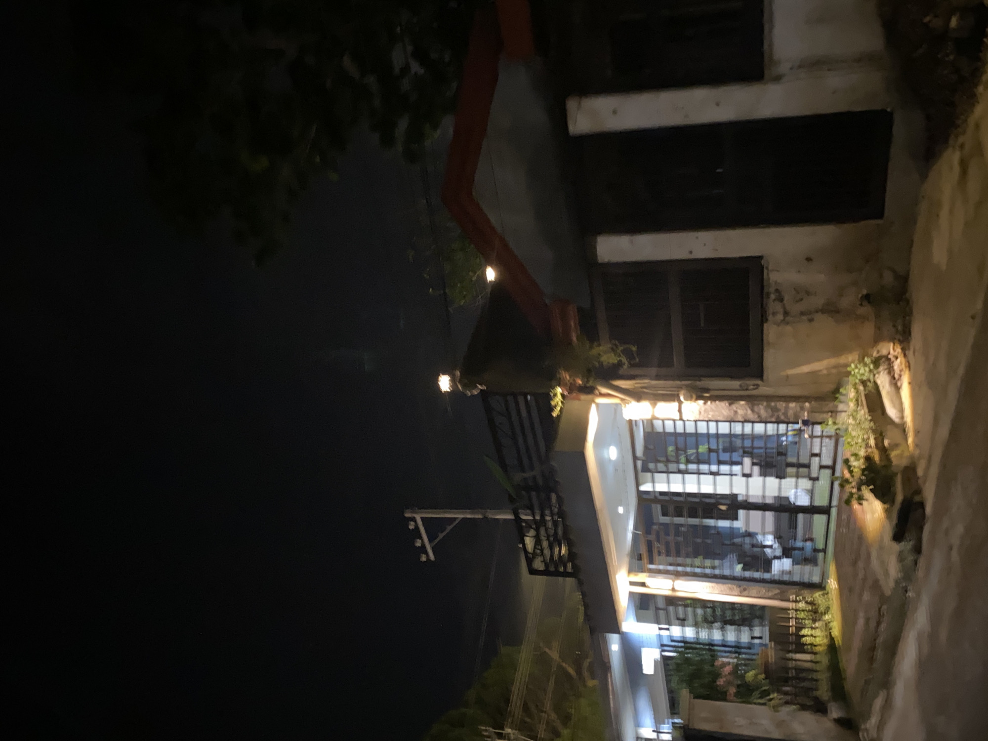 This nighttime image depicts an urban residential street in Davao City, Philippines. The scene features two distinct buildings illuminated by artificial light against a dark sky with faint atmospheric haze.
On the left, a modern multi-story structure presents white walls, a dark metal balcony railing on its upper level, and a prominent dark metal gate at the entrance, characterized by a grid-like pattern of horizontal and vertical bars. The facade is extensively lit by multiple recessed lights under an overhang and vertical strip lights on its pillars. A small landscaped patch with green foliage is visible at ground level in front of the gate.
Adjacent to the right, a second building displays a contrasting architectural style with a distinct reddish-orange gabled roof. Its lower walls are light-colored and show signs of weathering. This structure features dark windows with grates and a dark door. Utility components, including an electrical meter, are visible on its exterior. String lights are affixed near its roofline, extending towards the left.
In the mid-ground, positioned between the buildings and behind the left structure's balcony, a concrete utility pole with power lines is present. Dark tree foliage is discernible on the far right and left edges of the frame. The foreground consists of a paved surface, indicative of a street or pathway, with small areas of vegetation and scattered lighting from the buildings. No individuals are present.