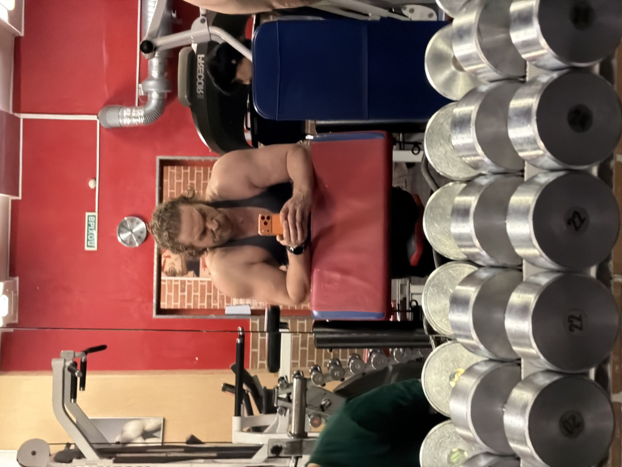The image shows a man with fair skin and curly brown hair taking a selfie in a gym in Saint Petersburg, Russia. He is wearing a dark gray tank top and a smartwatch on his left wrist. The man is sitting on a workout bench with a red seat and a blue back. He is holding an orange-colored smartphone. To the right of the bench are rows of silver dumbbells, with the number "22" visible on some of them. Behind the man, there's a red wall with a framed picture and a clock. Further back, there's gym equipment, including a rowing machine. The lighting appears to be artificial, and the overall environment suggests a typical gym setting.