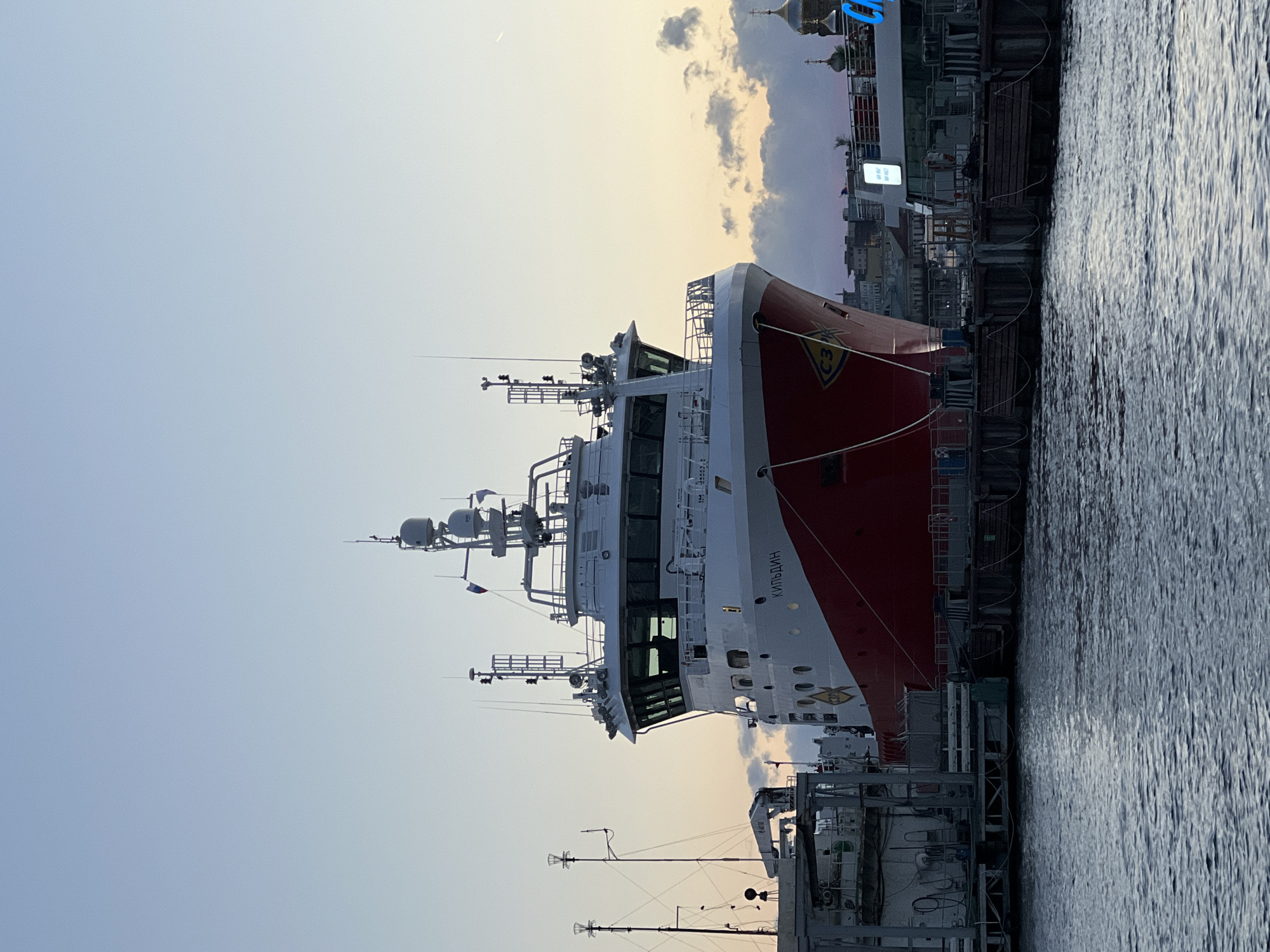 The image shows the Russian research vessel "Kildin" docked in Saint Petersburg, Russia. The ship is primarily red and white. The bridge of the ship faces the viewer. It is tied to a dock on the right side of the image. In the background are buildings, including a domed structure, that are consistent with the city's architecture. The sky is light, suggesting either early morning or late evening. The water in the foreground is dark. No people are visible in the image.