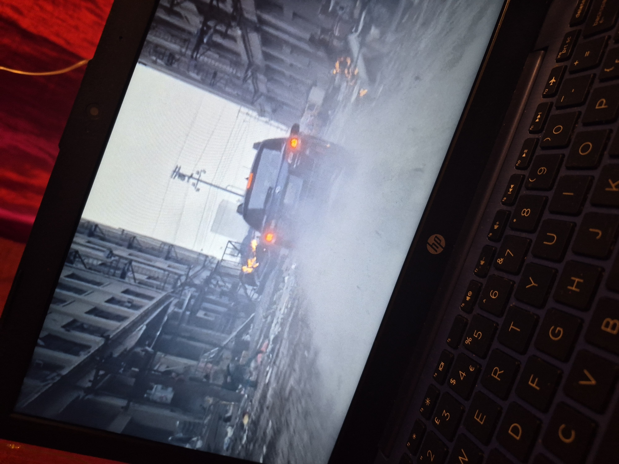 The image shows a close-up, angled view of a laptop screen displaying a scene from what appears to be a video game or movie. The screen content depicts a dark, possibly urban environment with a vehicle in motion, creating a significant amount of dust or smoke. The vehicle's taillights are illuminated and appear red and orange, suggesting brake lights or exhaust heat. The background of the screen shows the blurred outlines of buildings or structures, contributing to a sense of chaos or action.
The laptop itself is a dark color with a visible keyboard. The keys are illuminated by the screen's glow, and some keys are legible, including "hp" logo, function keys, and alphabet keys like "P," "Y," "R," "T," "U," "F," "G," "H," "J," "V," and "B." Symbols and numbers like "$," "€," "%," "4," "5," "6," "7," "8," and "9" are also visible on the keys. The overall lighting suggests it might be nighttime or a dimly lit indoor space.
The visible text from the screen and keyboard is limited and fragmented, primarily consisting of keyboard characters and symbols. The scene on the laptop screen evokes a feeling of urgency or intensity due to the moving vehicle and the dust cloud. The specific location within Sandwell, United Kingdom, is not discernible from the image content.