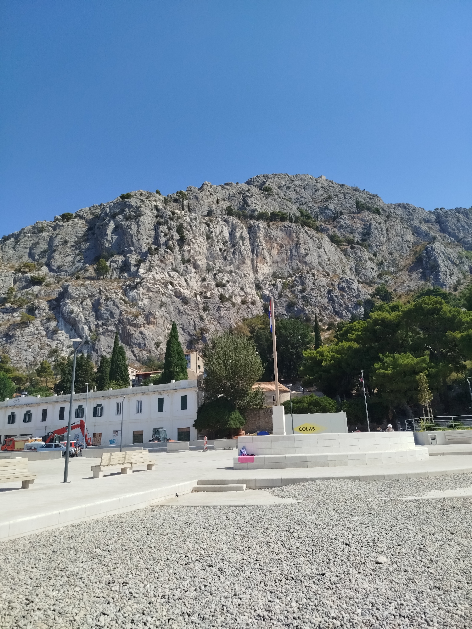 The image shows a wide view of a public square in Omiš, Croatia, with a prominent rocky mountain in the background. The square is paved with light-colored gravel in the foreground and concrete in the middle ground. Several modern, minimalist benches are placed on the concrete area.
In the midground, there is a long, white building with dark-windowed openings. In front of the building and to the right, a modern, multi-tiered white structure is visible, resembling a stage or seating area. A flag pole with a Croatian flag is mounted on this structure, along with a small, traditional-looking building with a red roof to its left. A yellow and white sign with the word "COLAS" is attached to the white structure.
Several green trees are scattered throughout the scene, with a dense cluster of dark green trees behind the white building and climbing up the lower slopes of the mountain. The mountain itself is a large, imposing rock formation with visible texture and crevices, largely devoid of vegetation except for sparse green patches. The sky is a clear, bright blue.
A red construction vehicle (excavator) and a white vehicle are parked near the white building. In the far right of the image, a few people are visible in the distance, appearing as small figures in the outdoor setting. The overall scene suggests a public space designed for recreation or events, set against a dramatic natural backdrop.
