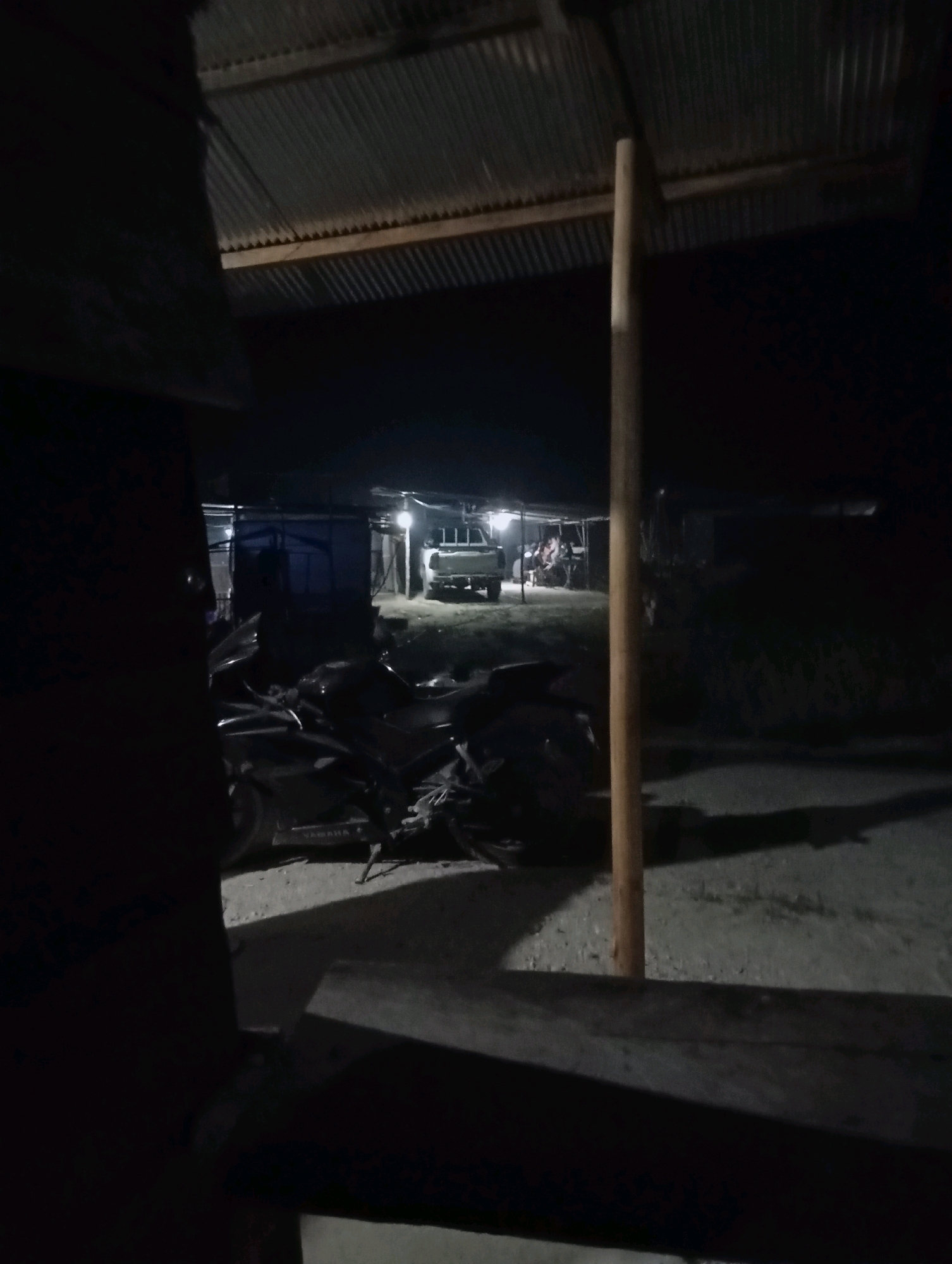 The image depicts a dimly lit outdoor scene at night in Tumbang Tariak, Indonesia. In the foreground, a dark-colored motorcycle is parked. To the right of the motorcycle, a vertical wooden pole stands. Behind the motorcycle, a corrugated metal roof supported by wooden beams is visible. Further in the background, a light-colored pickup truck is parked under a sheltered area, with several individuals gathered around it. The ground appears to be dirt or gravel, and shadows are cast across the scene, suggesting artificial lighting from the sheltered area.