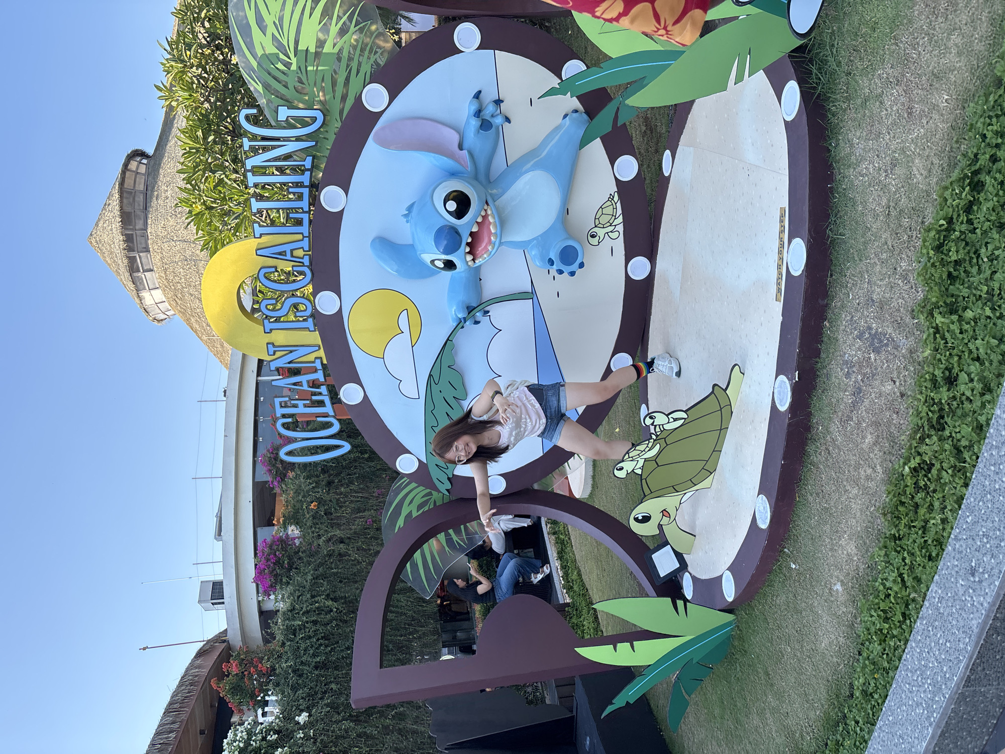 A young woman with glasses and long brown hair is posing for a photo in front of a large, circular display featuring Stitch and turtles, with the text "OCEAN IS CALLING" prominently displayed above. She is wearing a pink tank top, denim shorts, and white sneakers with colorful striped socks. She is leaning back and extending her right arm, as if interacting with the display. The display itself is a cutout with a tropical island scene, a smiling Stitch character, and green turtles. Behind the display, a building with a thatched roof and surrounding greenery are visible. In the background, two other people are seated on a bench, with one holding a phone. The scene appears to be an outdoor event or attraction in Kuta, Indonesia, given the tropical elements and the text on the display. The ground is covered with grass and a stone border.