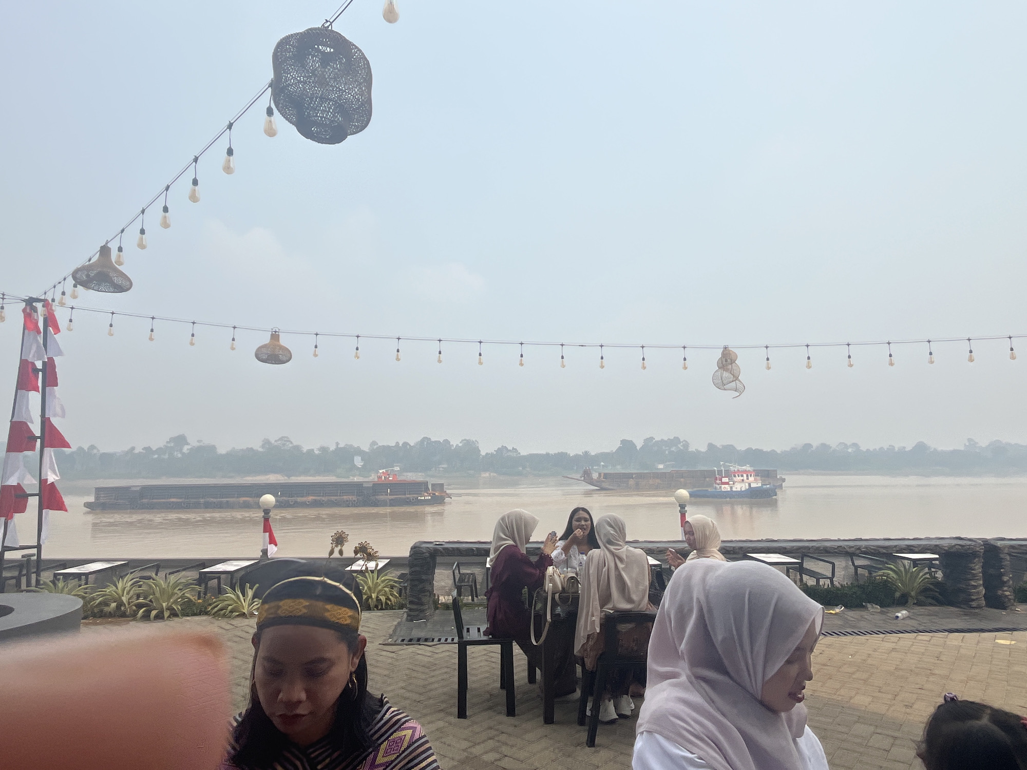This image captures an outdoor seating area overlooking a wide, calm river. The setting appears to be a cafe or restaurant along the riverbank in Meliau Hilir, Indonesia. In the foreground, a woman wearing a traditional head covering and a patterned top is seated, her gaze directed downwards. Several other individuals, mostly women dressed in hijabs, are seated at tables in the mid-ground, some engaged in conversation and others looking at their phones.
The river itself is a prominent feature, with two large barges or cargo ships navigating its waters. The barge on the left is much longer and appears to be transporting goods. The barge on the right is smaller and seems to be a tugboat or similar vessel, with a red and white superstructure. The riverbanks are lined with trees and vegetation, suggesting a natural, perhaps slightly hazy, environment.
Overhead, strings of lights and decorative woven fixtures are suspended, indicating an evening or overcast day setting designed for ambiance. A flagpole with Indonesian flags is also visible, reinforcing the location. The ground is paved with brick-like tiles. The overall atmosphere suggests a leisurely gathering place, possibly a popular spot for locals and visitors to enjoy the river views and socialize.