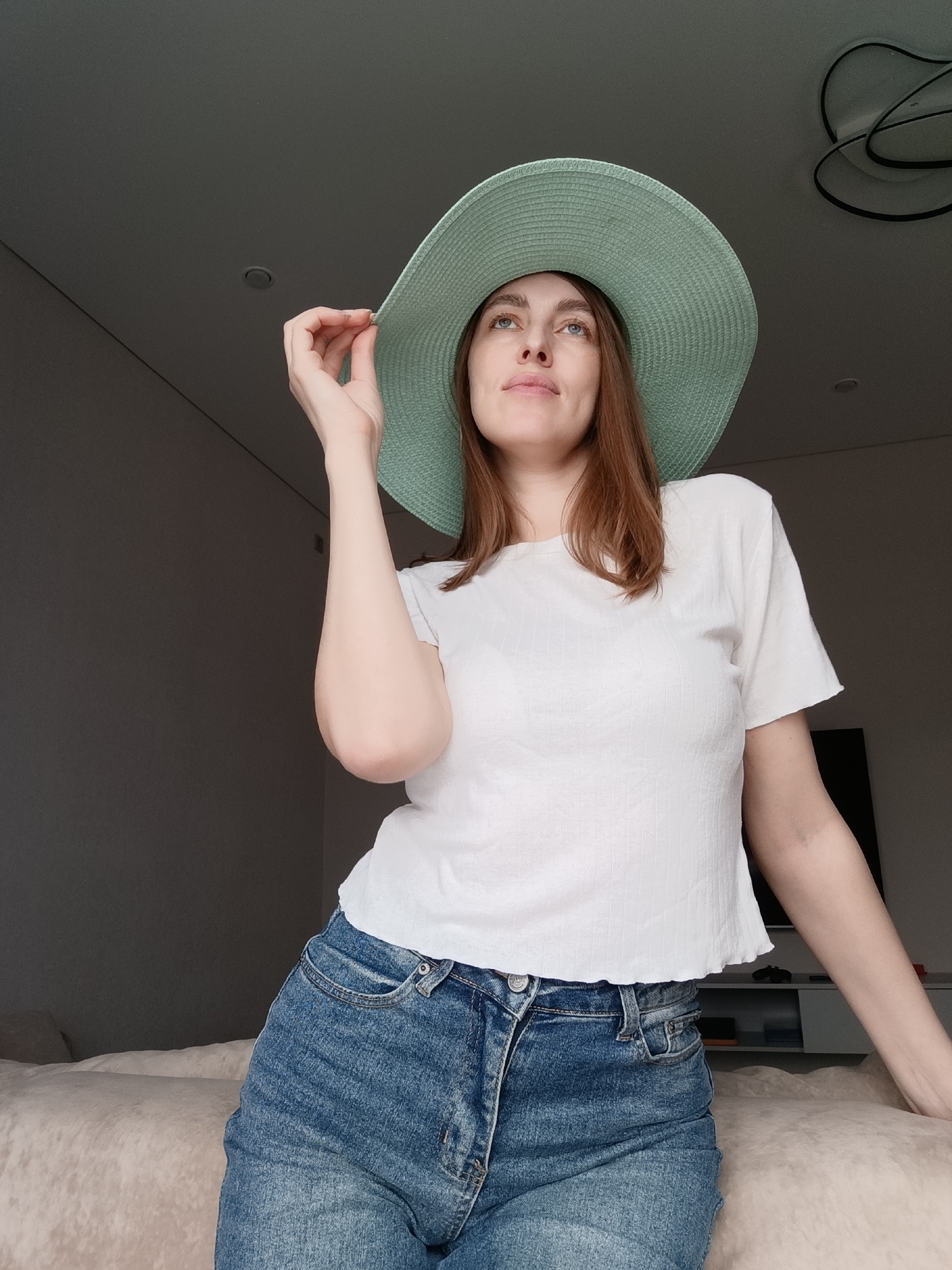 A young woman is the subject of this photograph, taken indoors, in what is reported to be Chaikovsky, Russia. She is wearing a wide-brimmed, light-green hat, a white short-sleeved shirt, and blue denim shorts. She holds the brim of her hat with her right hand and gazes upward and slightly to the right. Her hair is auburn and shoulder-length. The background includes neutral-toned walls, a bed with a light-colored covering, and a television with a modern entertainment unit beneath it. The lighting is diffused, and the overall atmosphere is relaxed and casual.