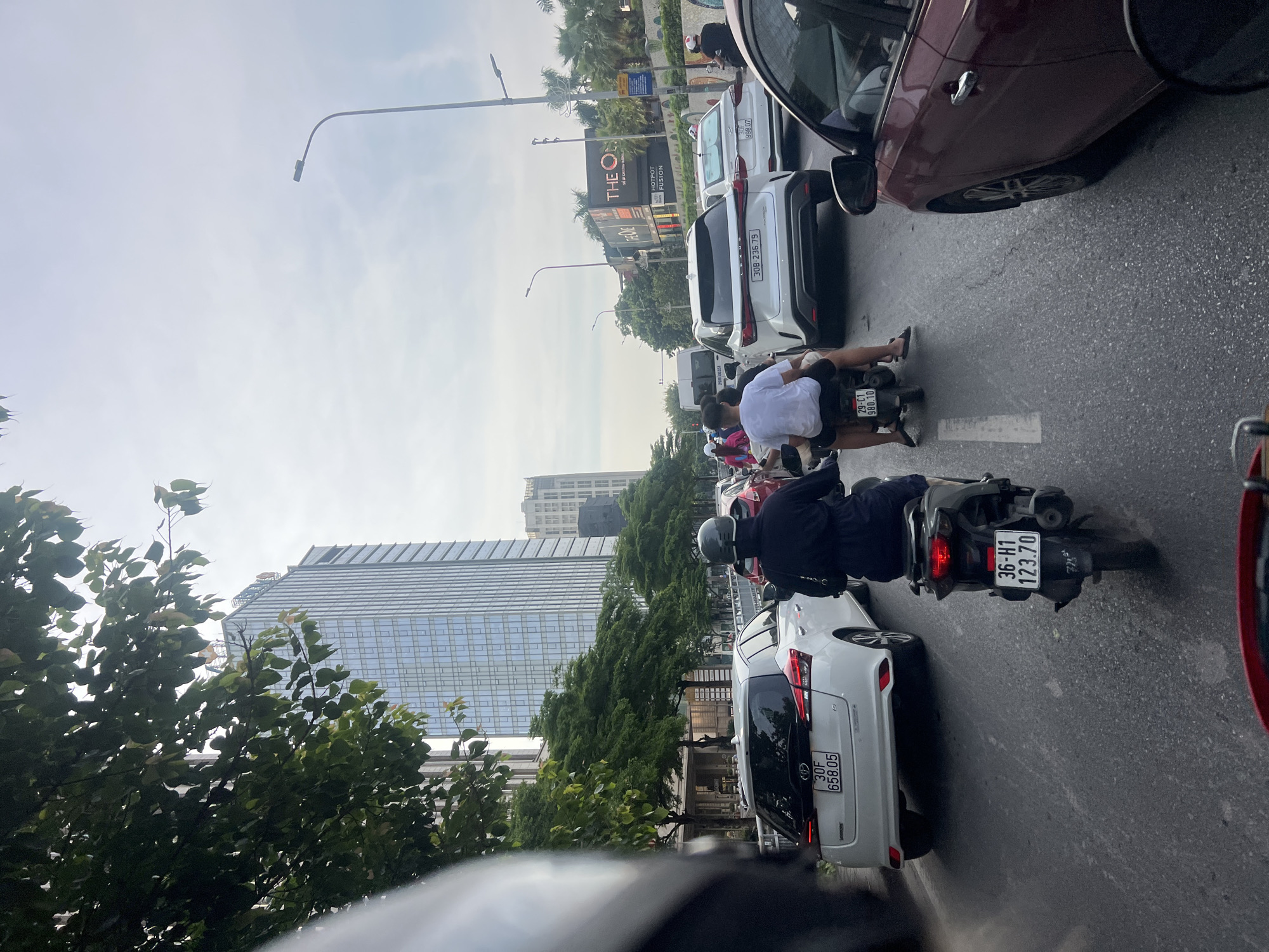 The image depicts a congested street scene in Hanoi, Vietnam, likely during daylight hours given the sky's appearance. The perspective is from a rider on a motorcycle, looking forward through a traffic jam.
In the foreground, the back of a black motorcycle is visible, with its license plate displaying "36-H1 123.70". A person in a black outfit, including a helmet, is seated on this motorcycle, facing forward. To the left of this motorcycle, the rear of a white hatchback car is prominent, with a license plate reading "30F 658.05".
Further ahead, several other vehicles are present, including a white Lexus SUV with a license plate "30B-236.79". Behind the Lexus, another white car with license plate "30F 998 07" is visible. On the right side of the road, a red car is partially visible.
The background reveals a bustling city environment with tall modern buildings, including a prominent glass-fronted skyscraper. Trees line the street, and streetlights are also visible. The sky is overcast, suggesting a cloudy day. The overall impression is of a typical urban traffic scene in Hanoi, characterized by dense vehicle presence and multi-story architecture.
